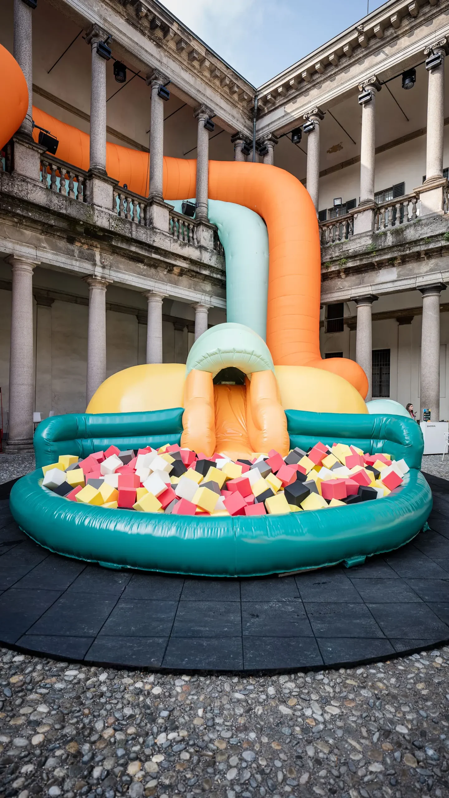 A large orange and mint-green inflatable slide leading into a teal ball pit filled with colorful foam cubes in the center of the courtyard.