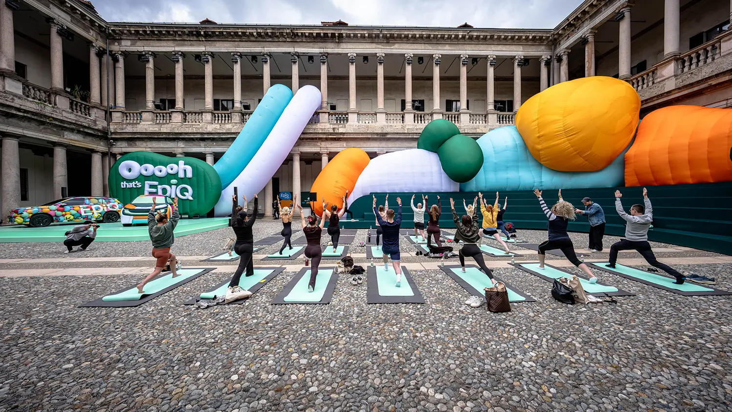 A group of people practicing yoga on teal mats in the center of the Palazzo del Senato courtyard, with large colorful inflatables in the background.