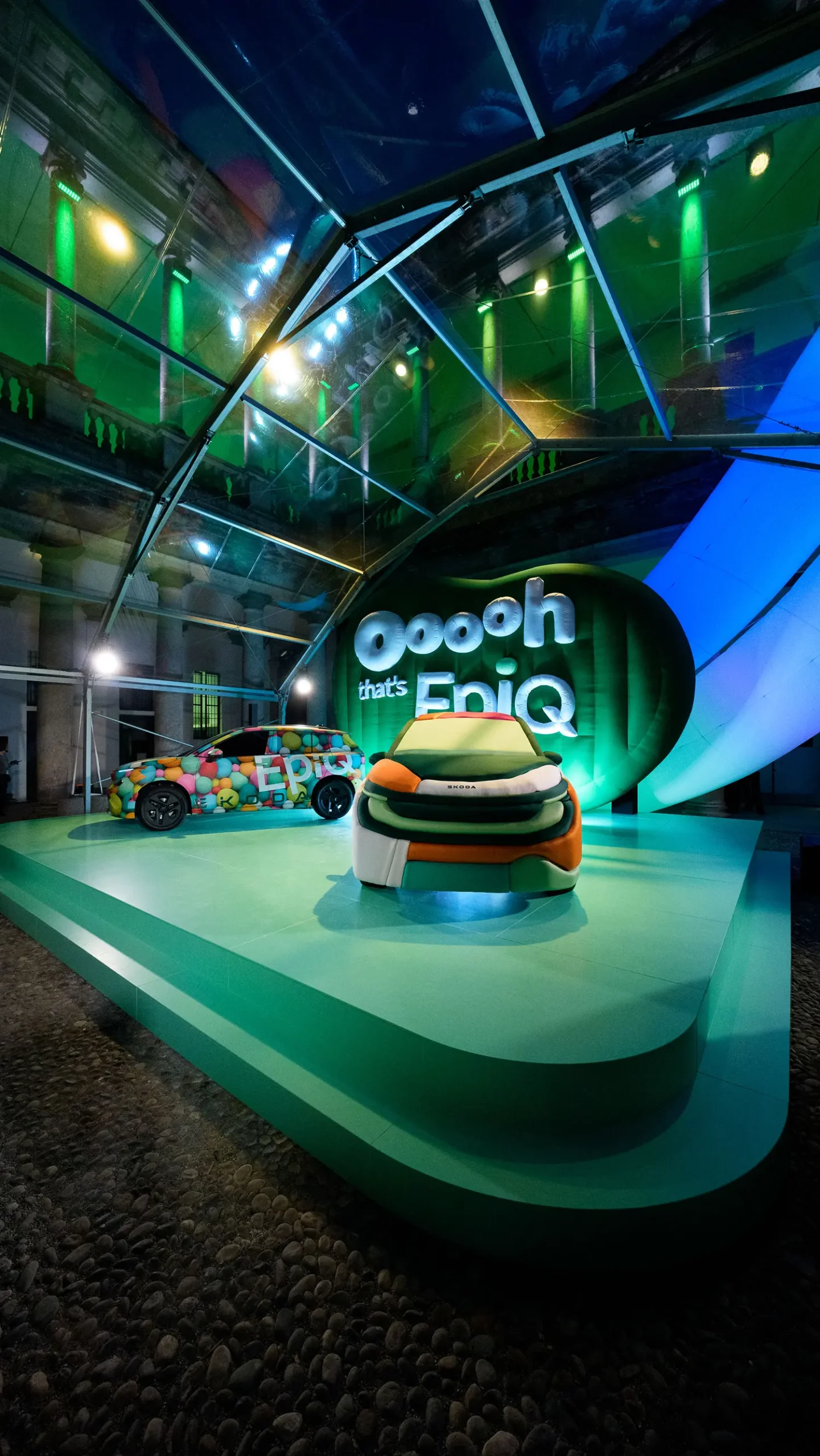 The Škoda Epiq Sculpt illuminated by green light at night, viewed through the glass structure of a modern pavilion in the courtyard.