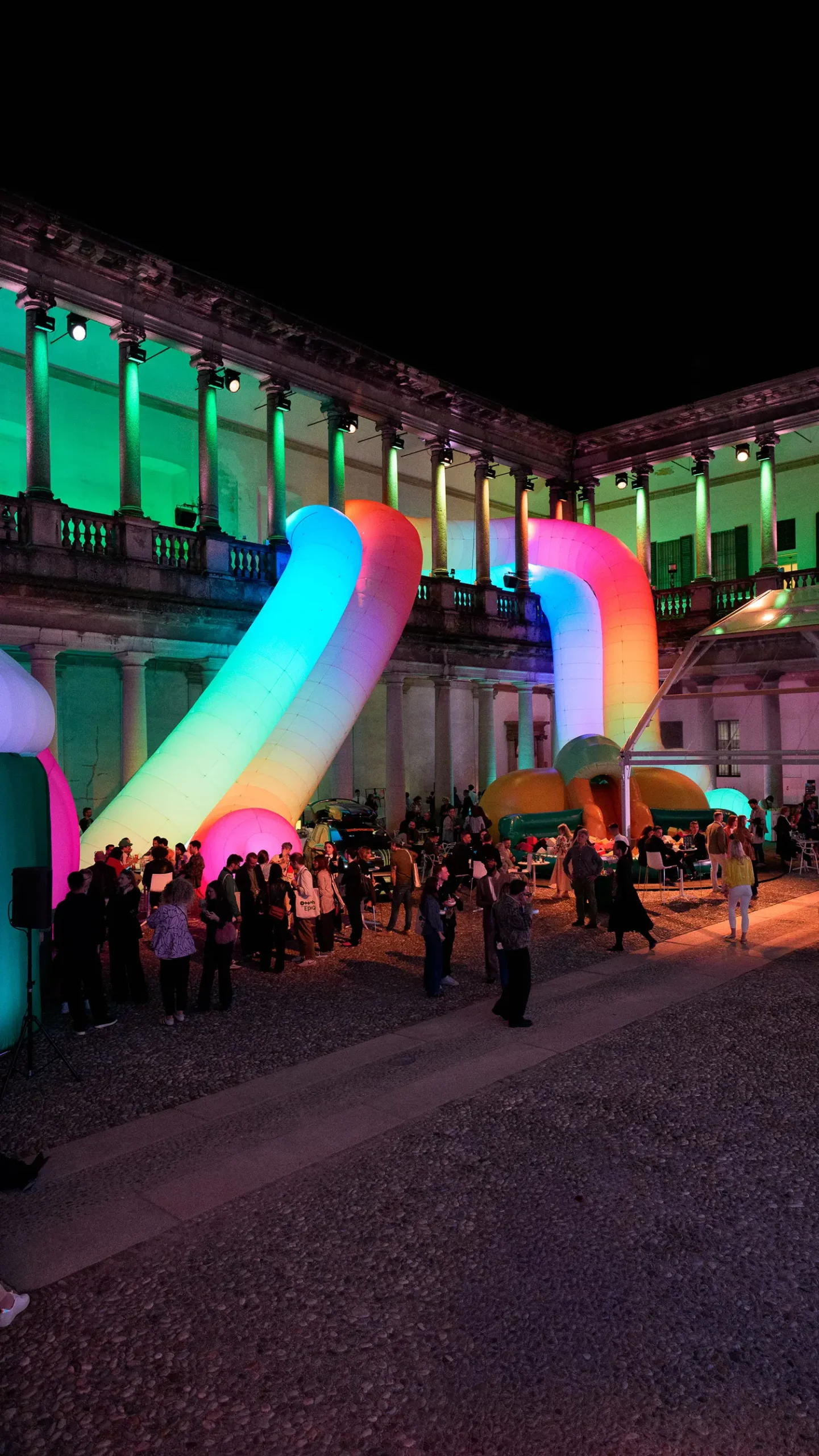 A night shot of the courtyard filled with people, featuring glowing inflatable tubes in blue, orange, and pink arching over the crowd.