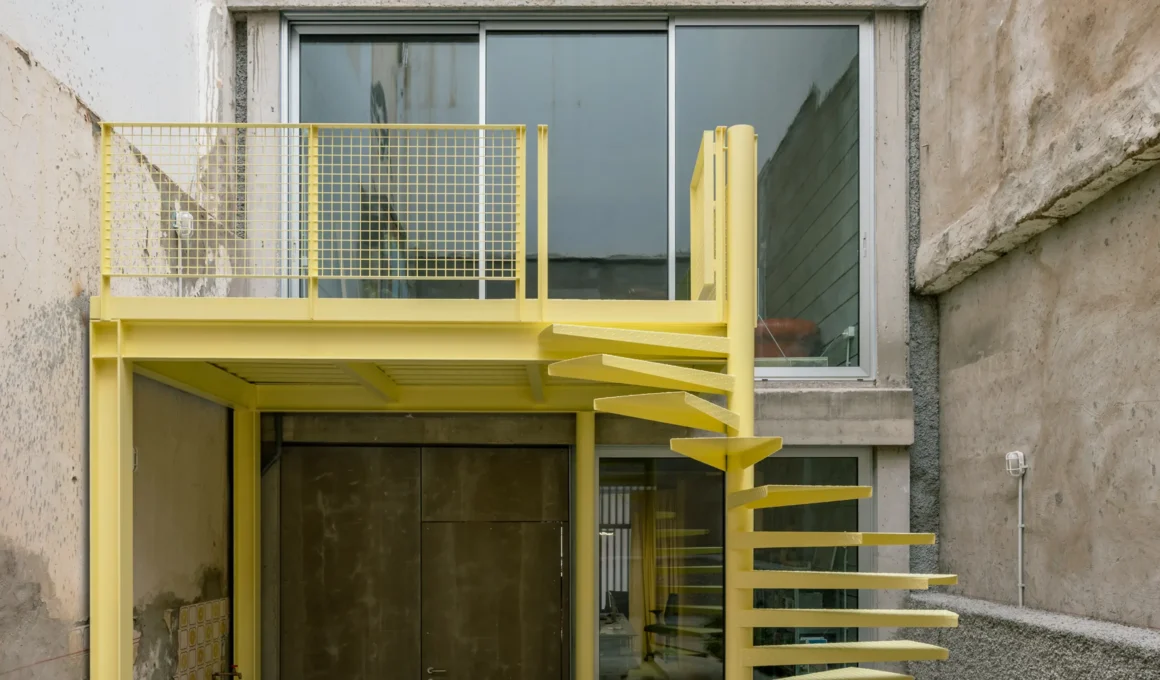 A vibrant yellow metal spiral staircase and elevated balcony against raw concrete walls in a courtyard.