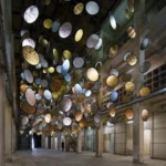 Wide view of HALOS installation by SpY in a tall industrial atrium with suspended gold and silver discs.