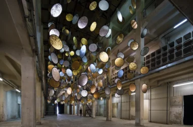 Wide view of HALOS installation by SpY in a tall industrial atrium with suspended gold and silver discs.
