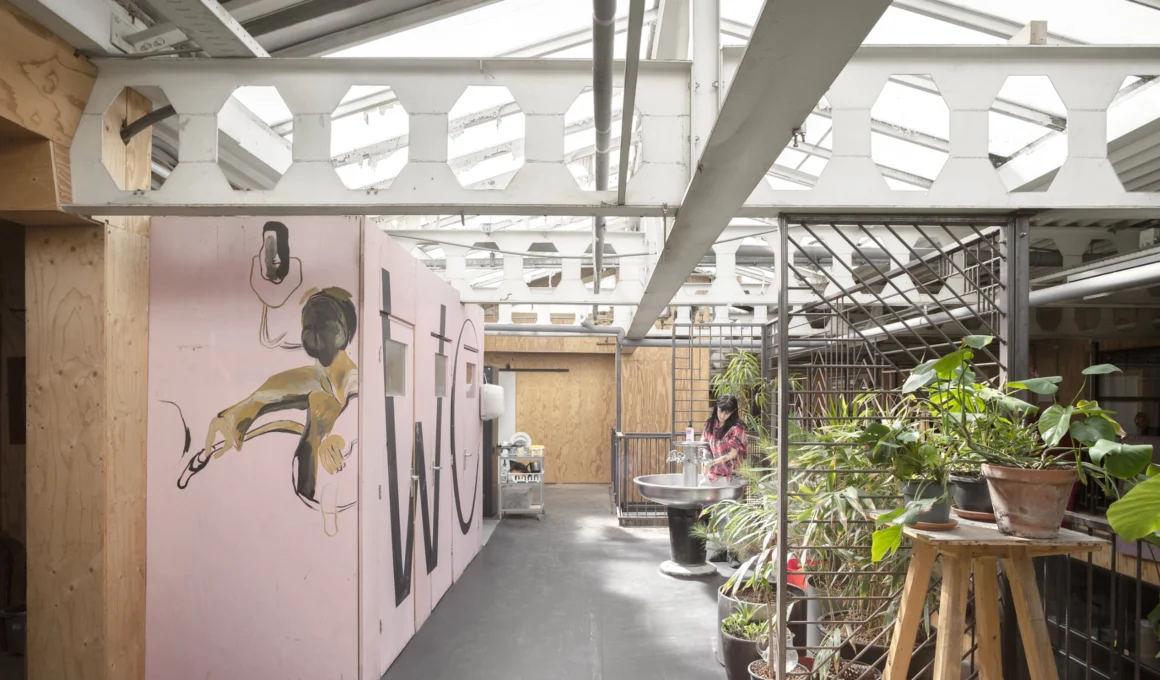A pink plywood structure with "WC" text and hand-painted figures under a sawtooth industrial roof.