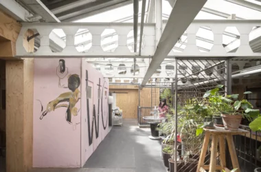 A pink plywood structure with "WC" text and hand-painted figures under a sawtooth industrial roof.