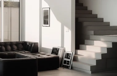 Contemporary living room showing visual weight with a black tufted sofa and a minimalist white concrete staircase under sharp natural light shadows.