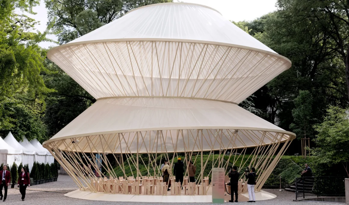 A wide-angle view of a two-tiered conical pavilion with a translucent white skin and thin wooden supports in a garden setting.