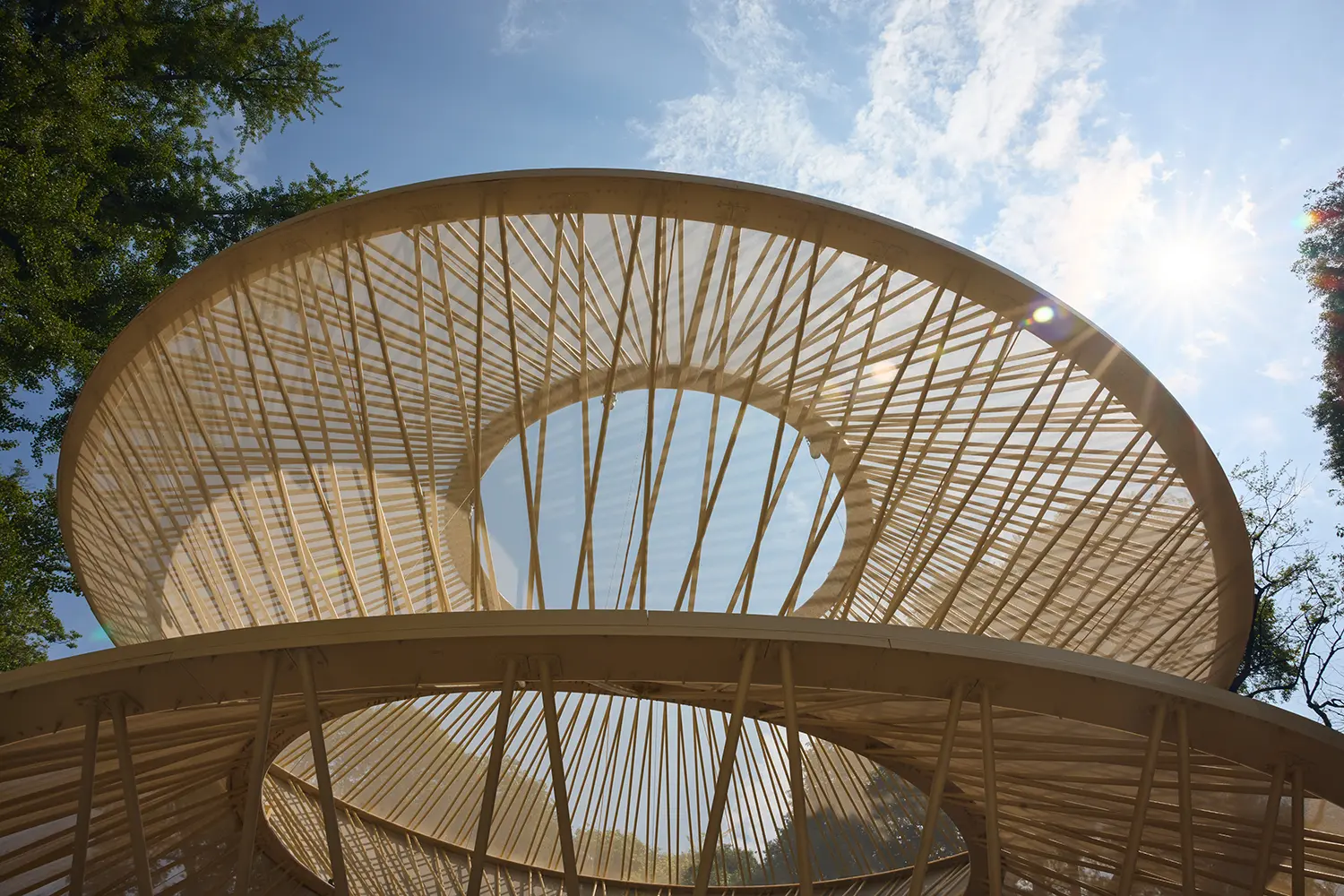 Low-angle perspective looking up at the radial wooden latticework of a circular pavilion roof against a bright blue sky.