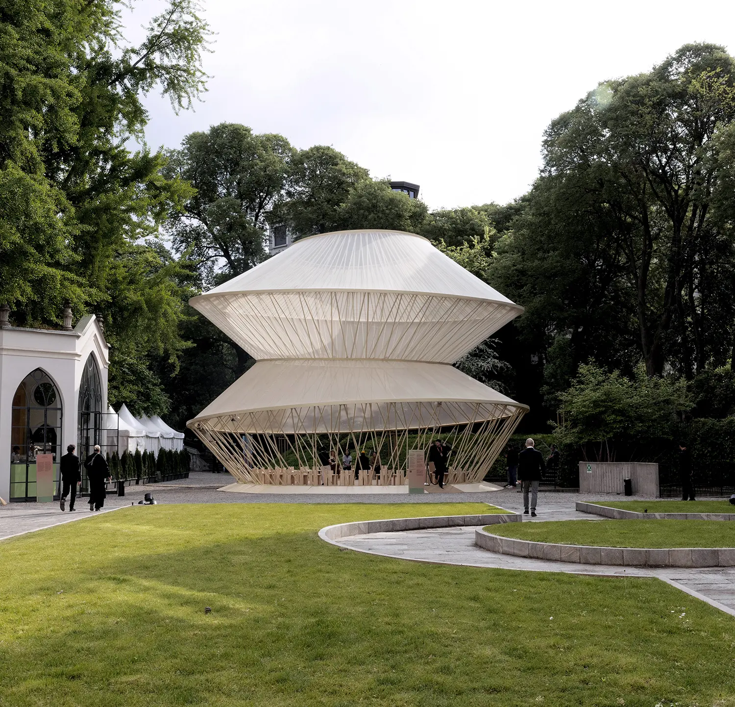 A distant view of the translucent tiered pavilion situated within the lush green context of a historic Italian garden.