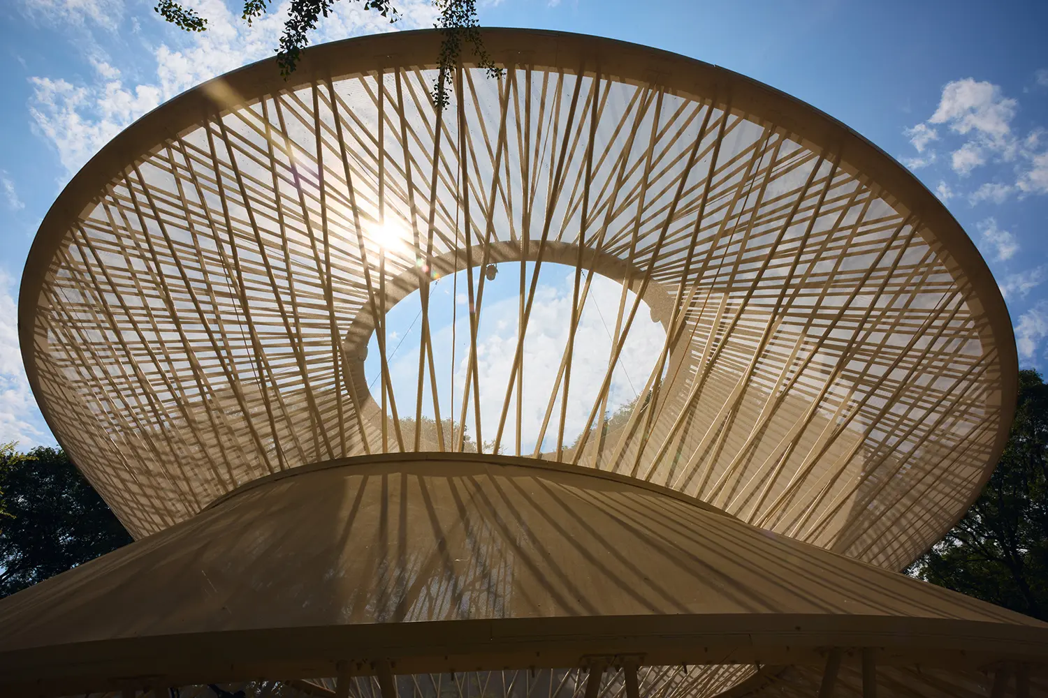 An upward view looking through the central oculus of a wooden pavilion, with the sun creating a lens flare effect.