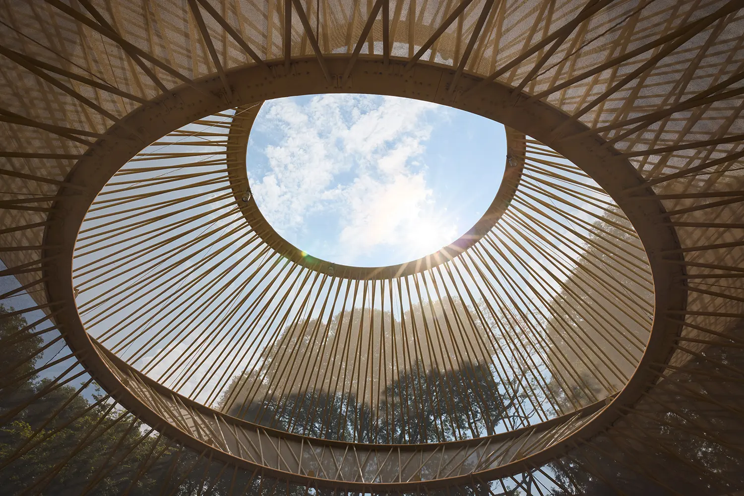 A vertical perspective from inside the pavilion looking through the circular top opening toward the sky and surrounding trees.