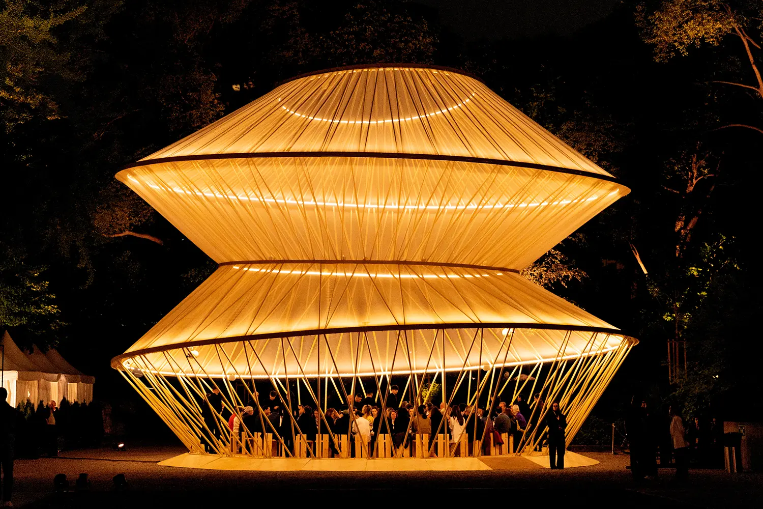 A night shot of the tiered pavilion glowing with warm internal light, with people gathered inside for an event.