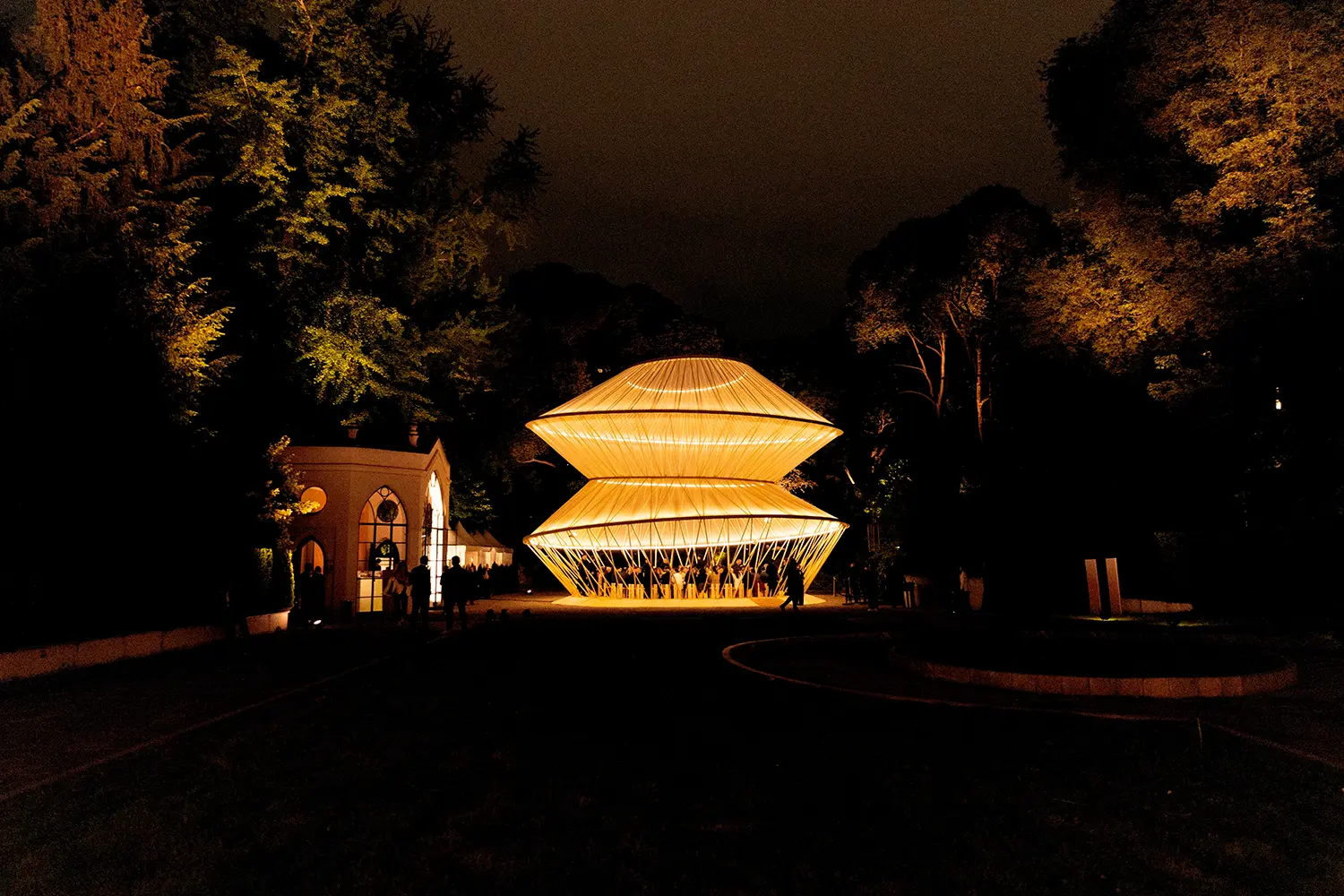 A long-distance night view of the illuminated pavilion as a central focal point in a dark garden landscape.