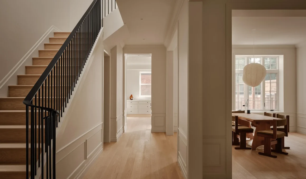 A long interior hallway in a Montreal home with white oak flooring, decorative wall moldings, and a black steel staircase railing on the left.