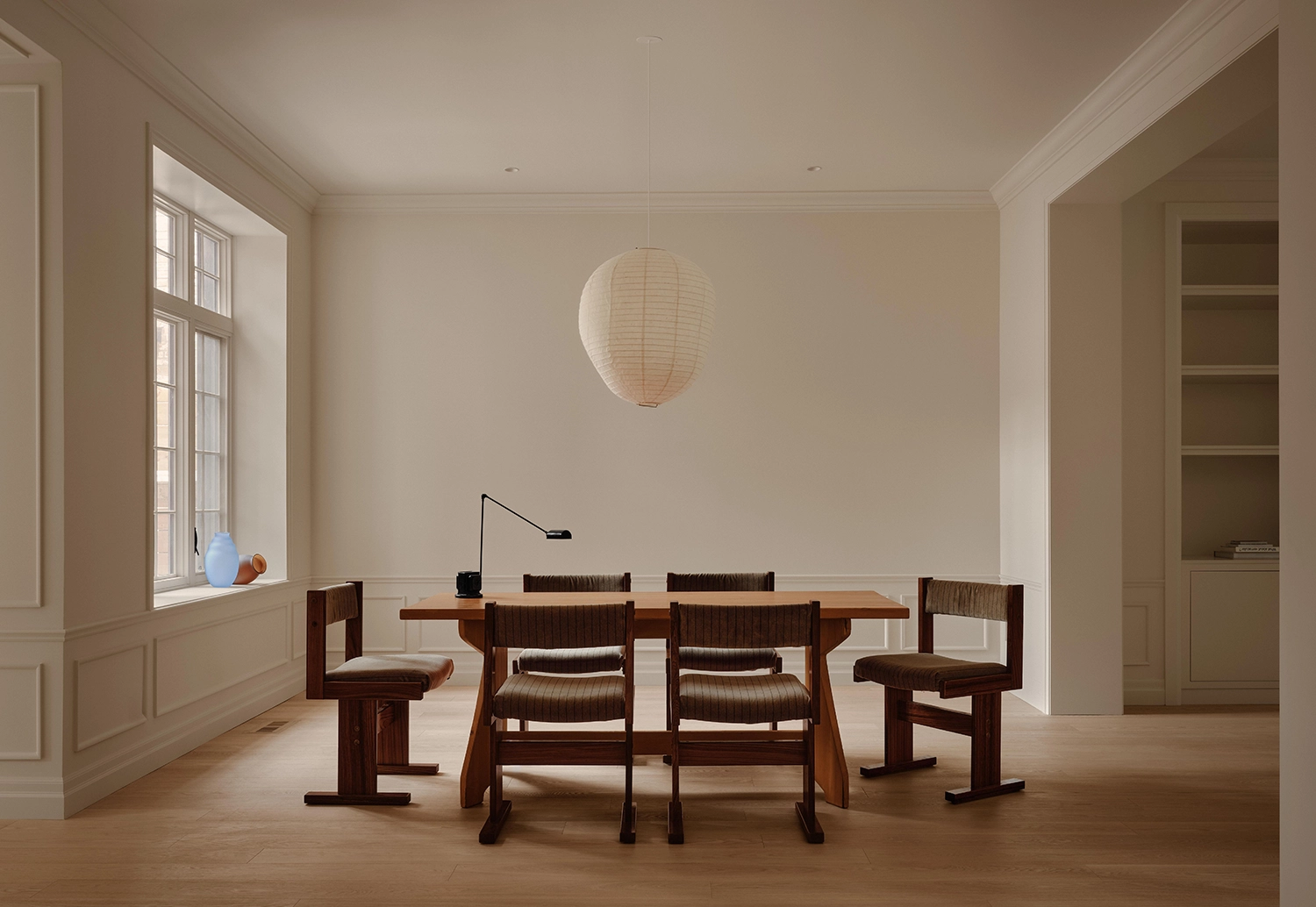 Symmetrical view of a minimalist dining room with a wooden table, four dark-toned chairs, a large paper lantern pendant, and a bright window.