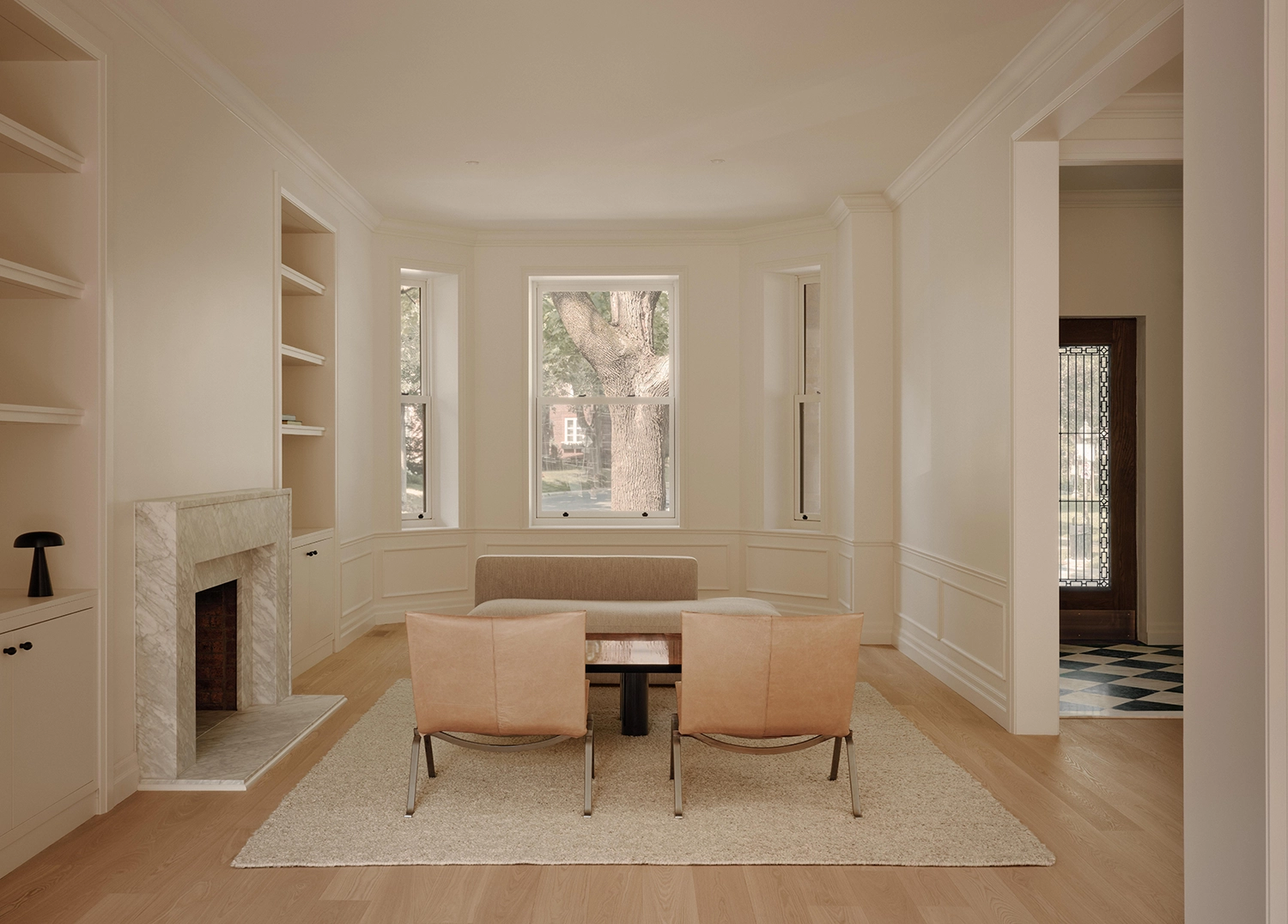 Wide view of a bright living room with a large bay window overlooking a tree, a marble fireplace, and neutral-colored contemporary furniture.
