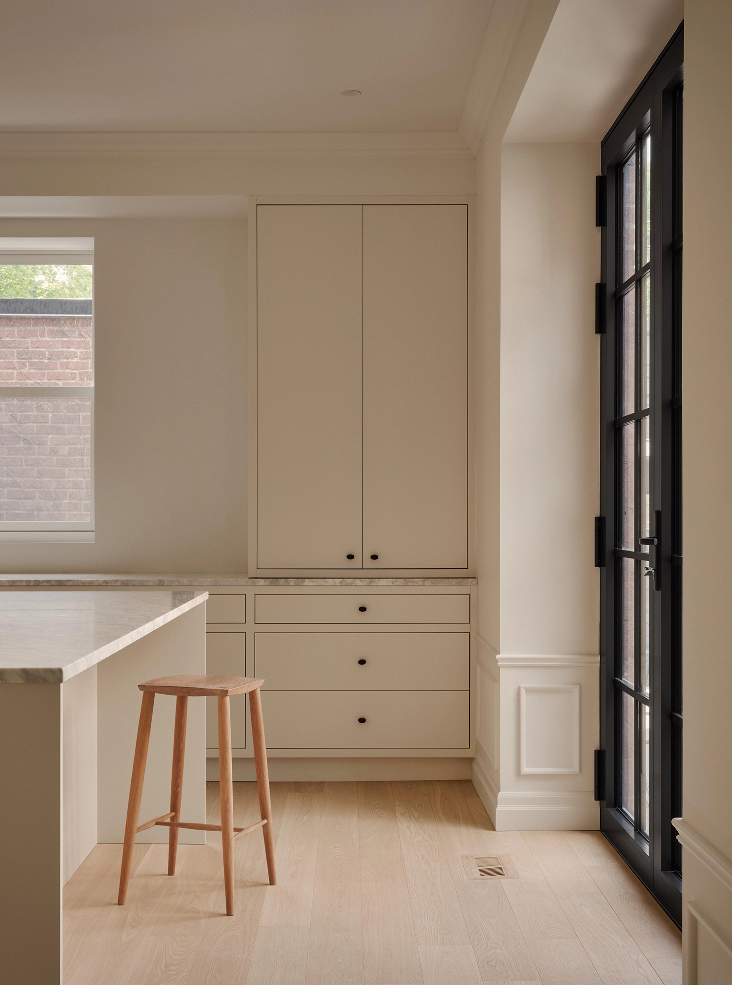 Close-up of a kitchen corner with a wooden stool, a white marble island, beige cabinetry, and a black-framed glass French door.