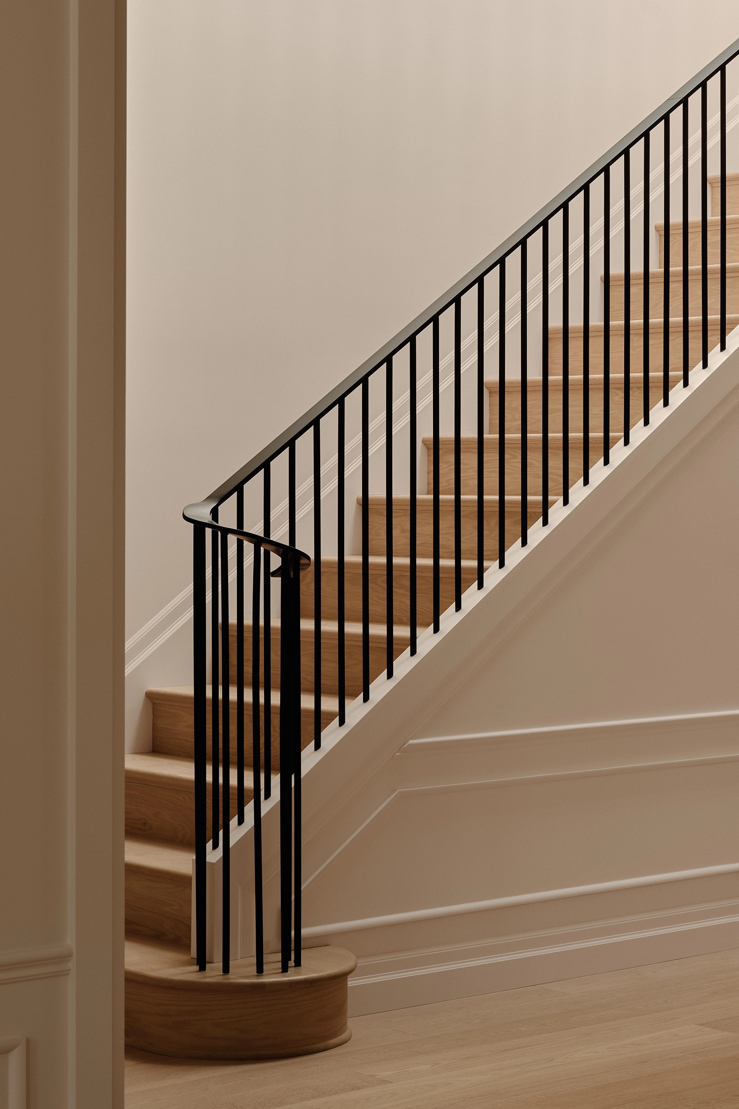 Detail shot of a staircase with light wood treads, a curved black steel handrail, and traditional white wall paneling.
