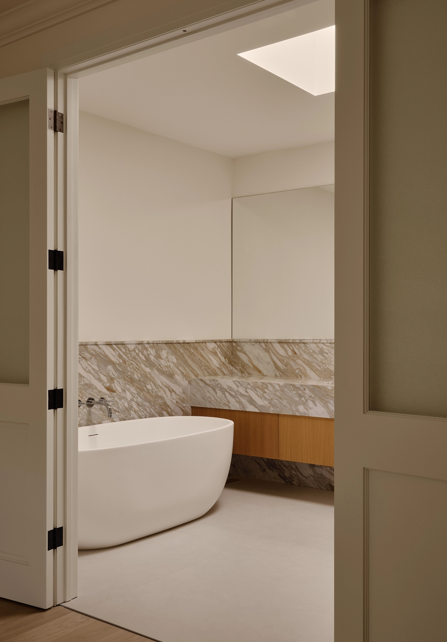 A modern bathroom viewed through open French doors, featuring a freestanding white bathtub and a thick marble vanity with a wooden base.