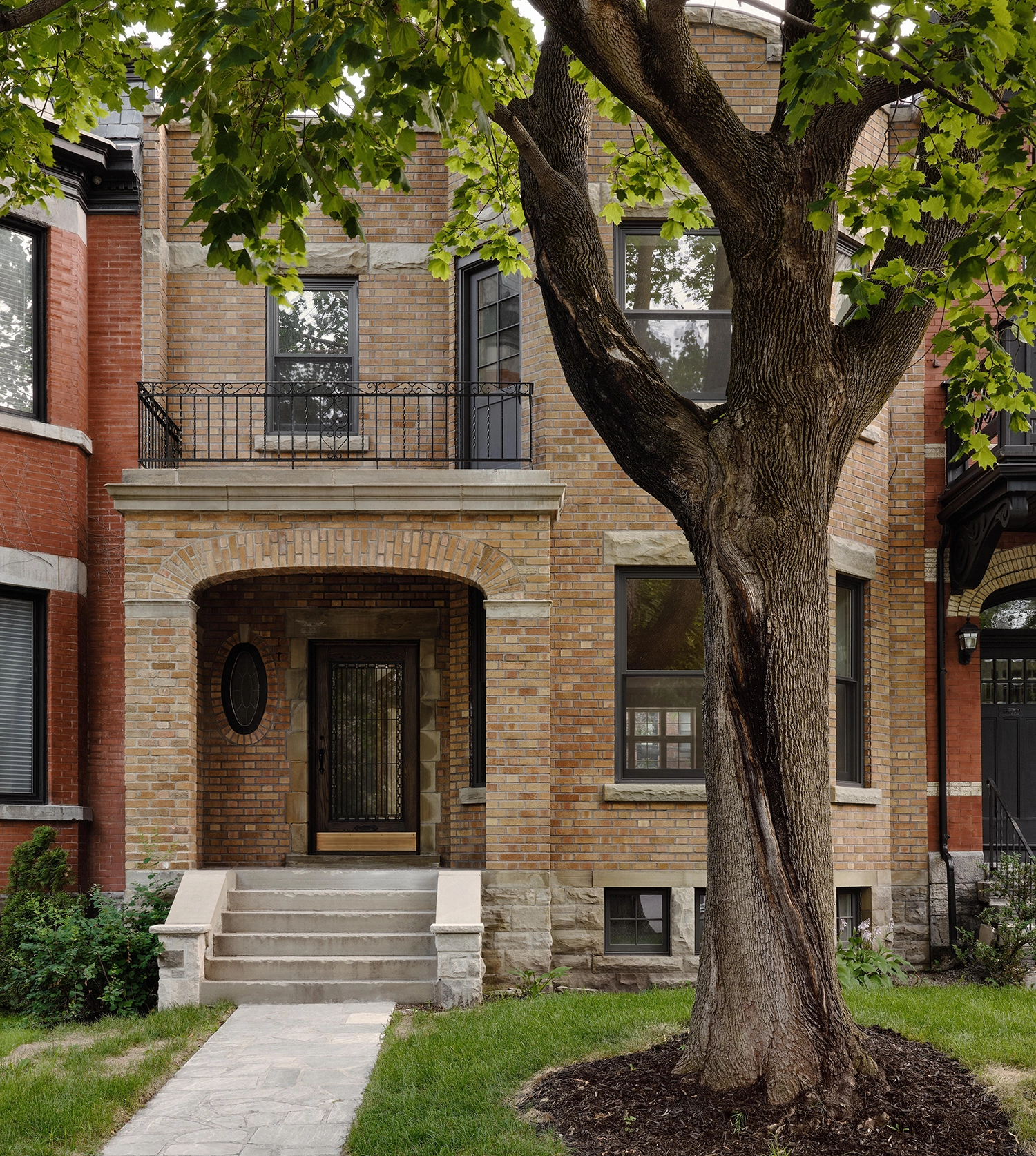 Exterior view of a traditional brick house in Montreal with a stone entrance, an oval window, and a large deciduous tree in the foreground.