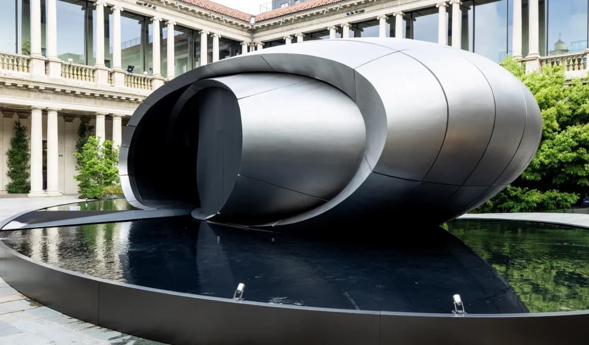 Exterior view of the sculptural Origin pavilion by Zaha Hadid Architects for Audi, featuring curved matt titanium panels over a dark reflecting pool.