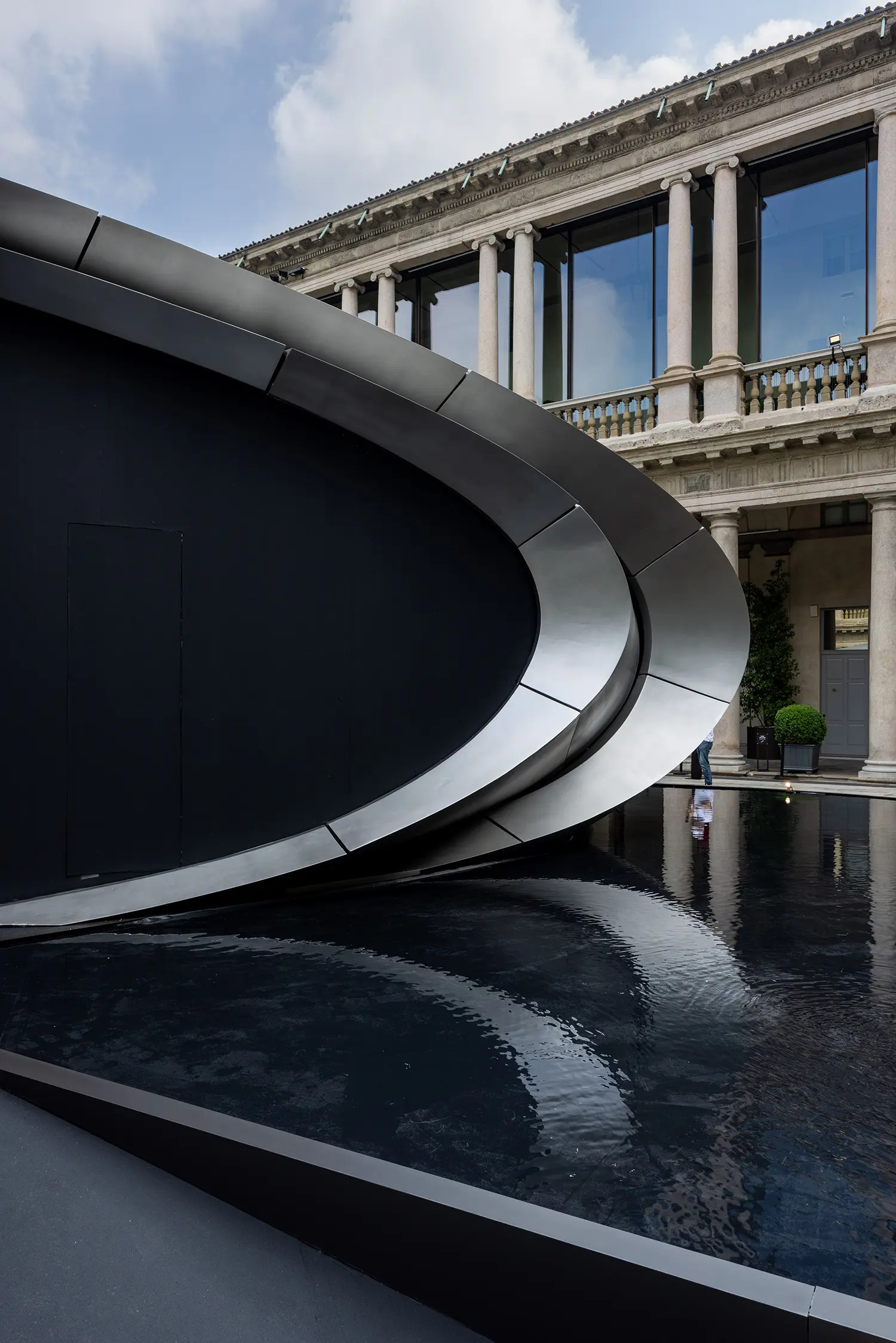 Close-up of the overlapping metallic layers of the Origin pavilion's entrance, showcasing sleek lines and the surrounding courtyard reflection.