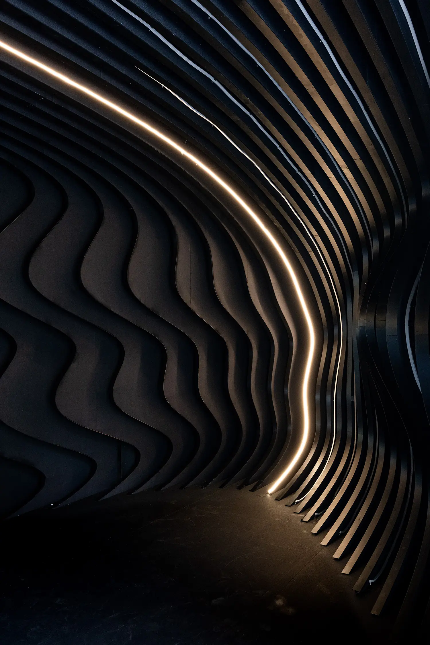Interior of the Origin pavilion showing dark, undulating wall ribs illuminated by a single, continuous line of warm LED light.