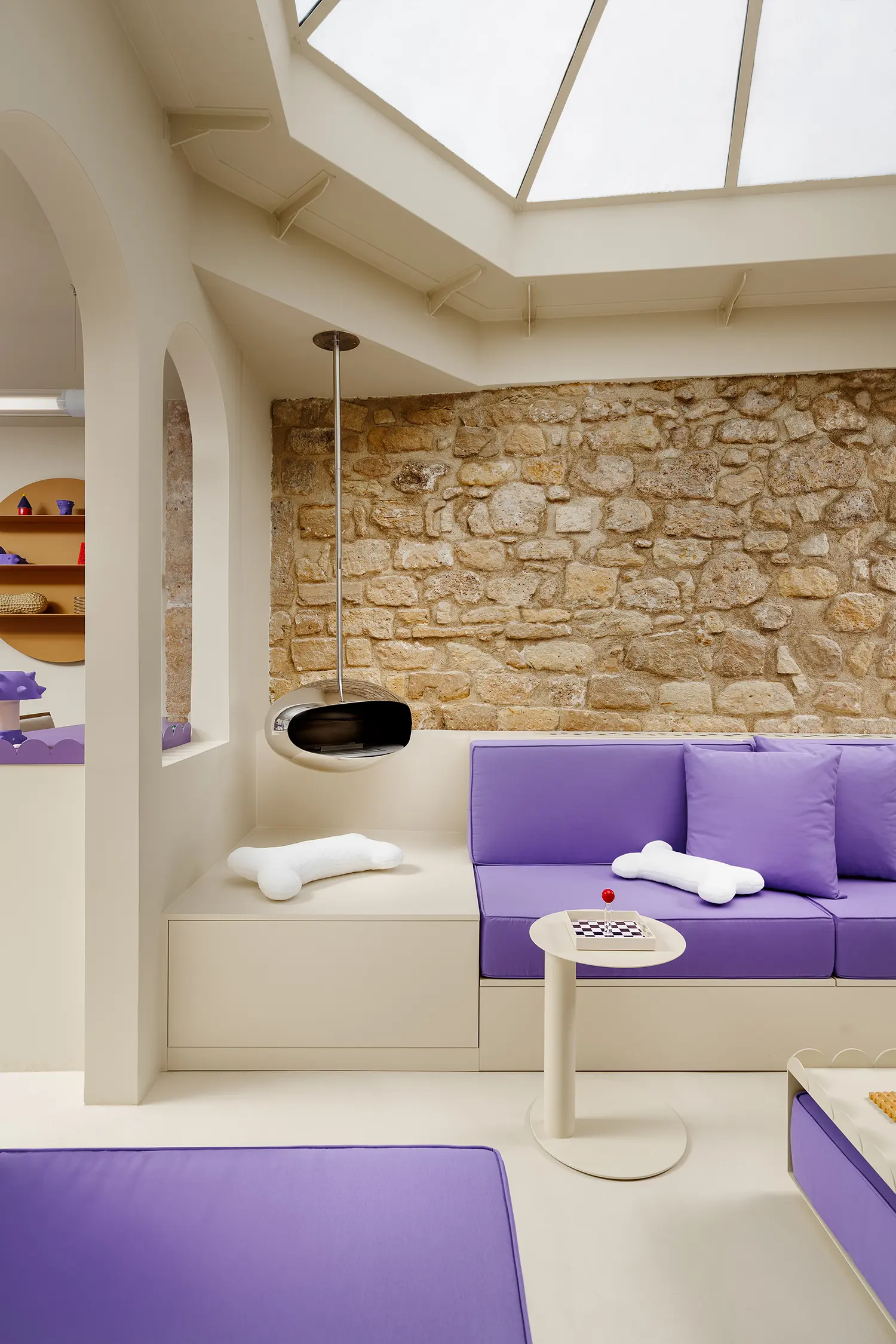 Chrome suspended fireplace next to an exposed stone wall and purple lounge seating under a skylight.