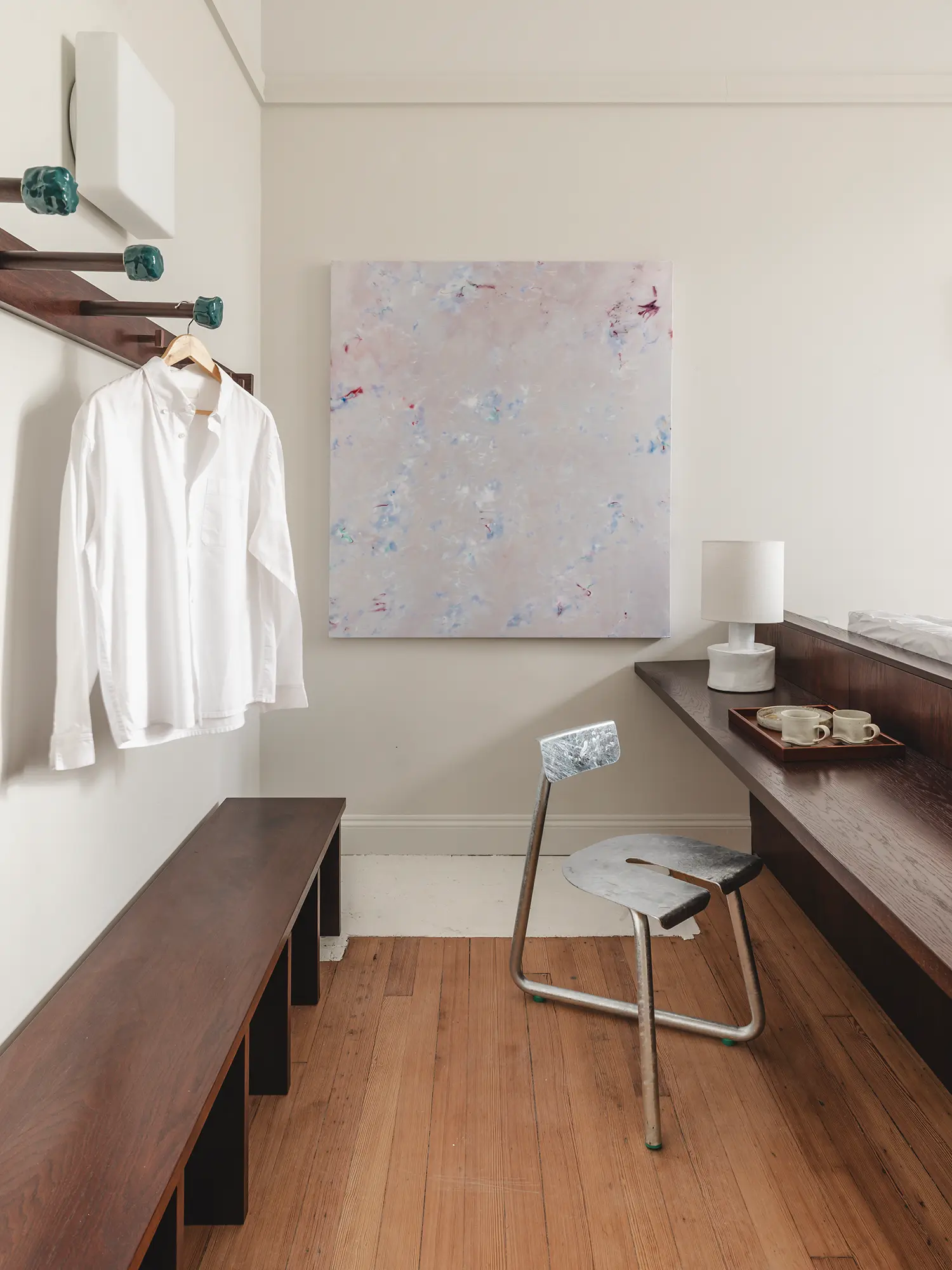 Minimalist hotel room corner with a wooden bench, industrial chair, and abstract painting.