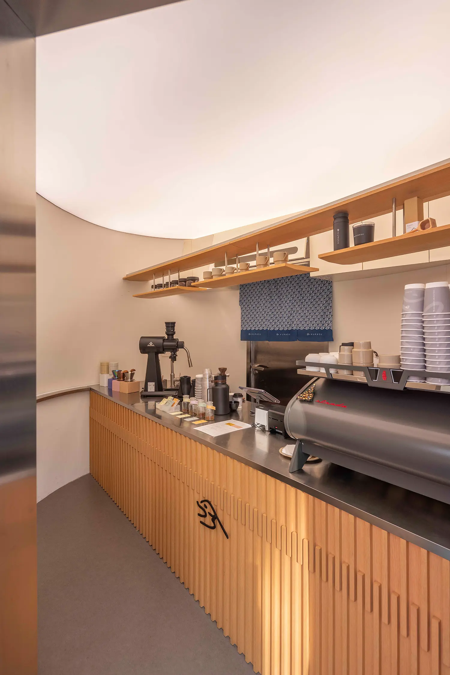 Interior view of the coffee bar featuring a slatted wooden counter, stainless steel equipment, and a glowing ceiling.