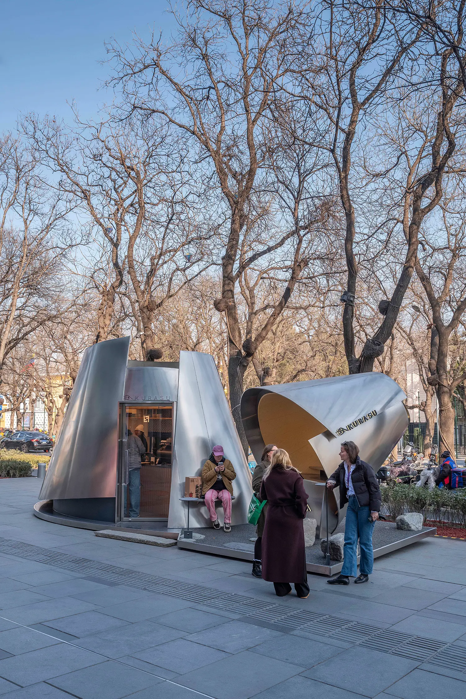 People gathered around the Kurasu coffee pavilion in a pedestrian plaza lined with winter trees.