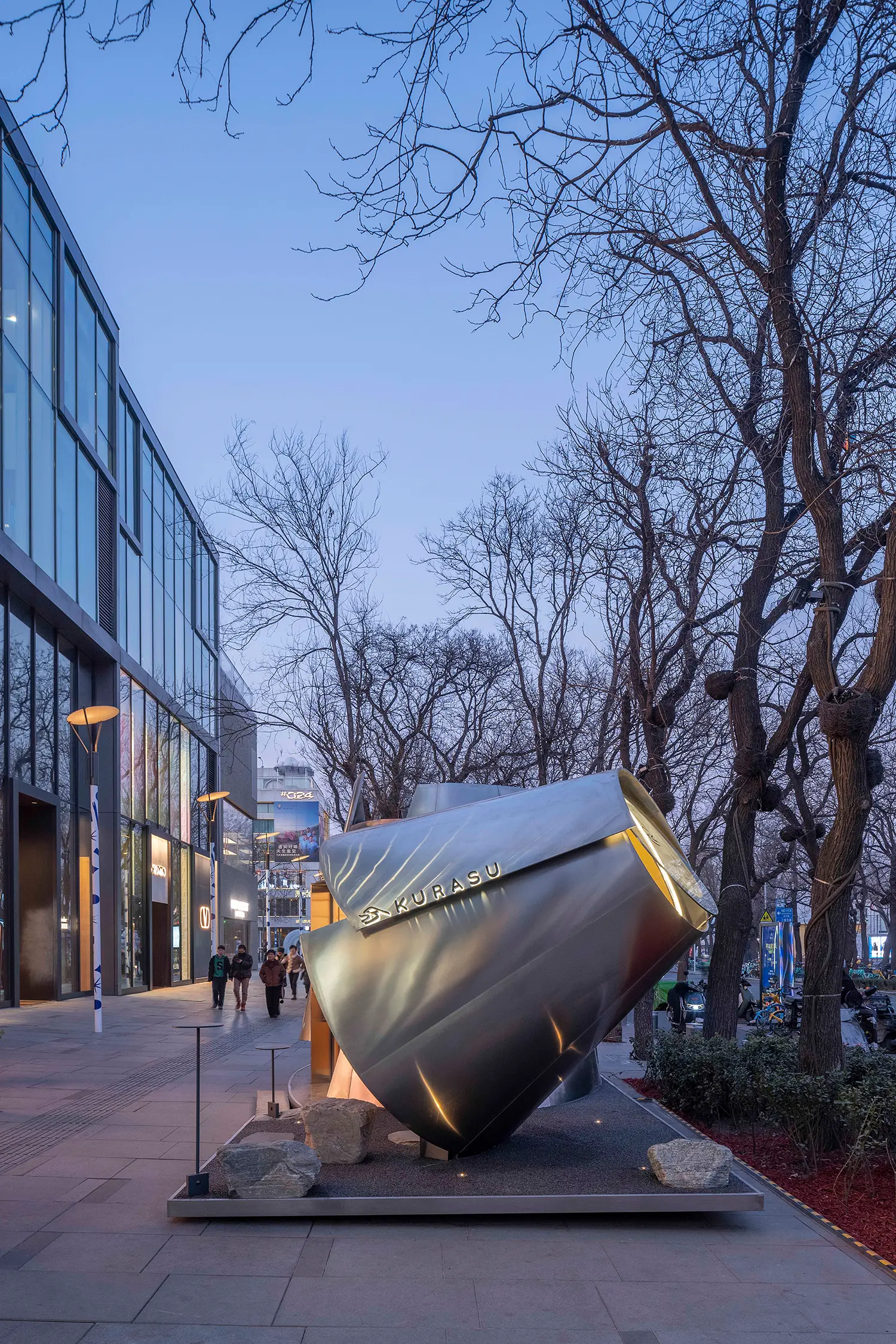 Night view of the Kurasu pavilion showing the illuminated logo and warm light emanating from the volumes.