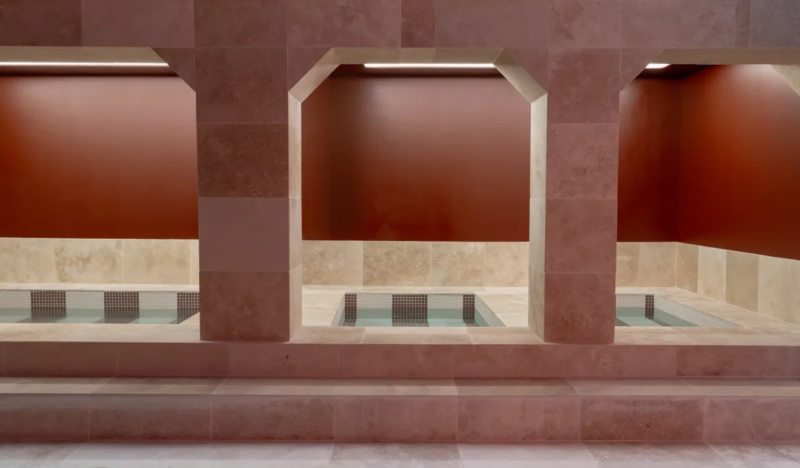 Three symmetric travertine archways overlooking square plunge pools with mosaic tiling and deep ochre walls.