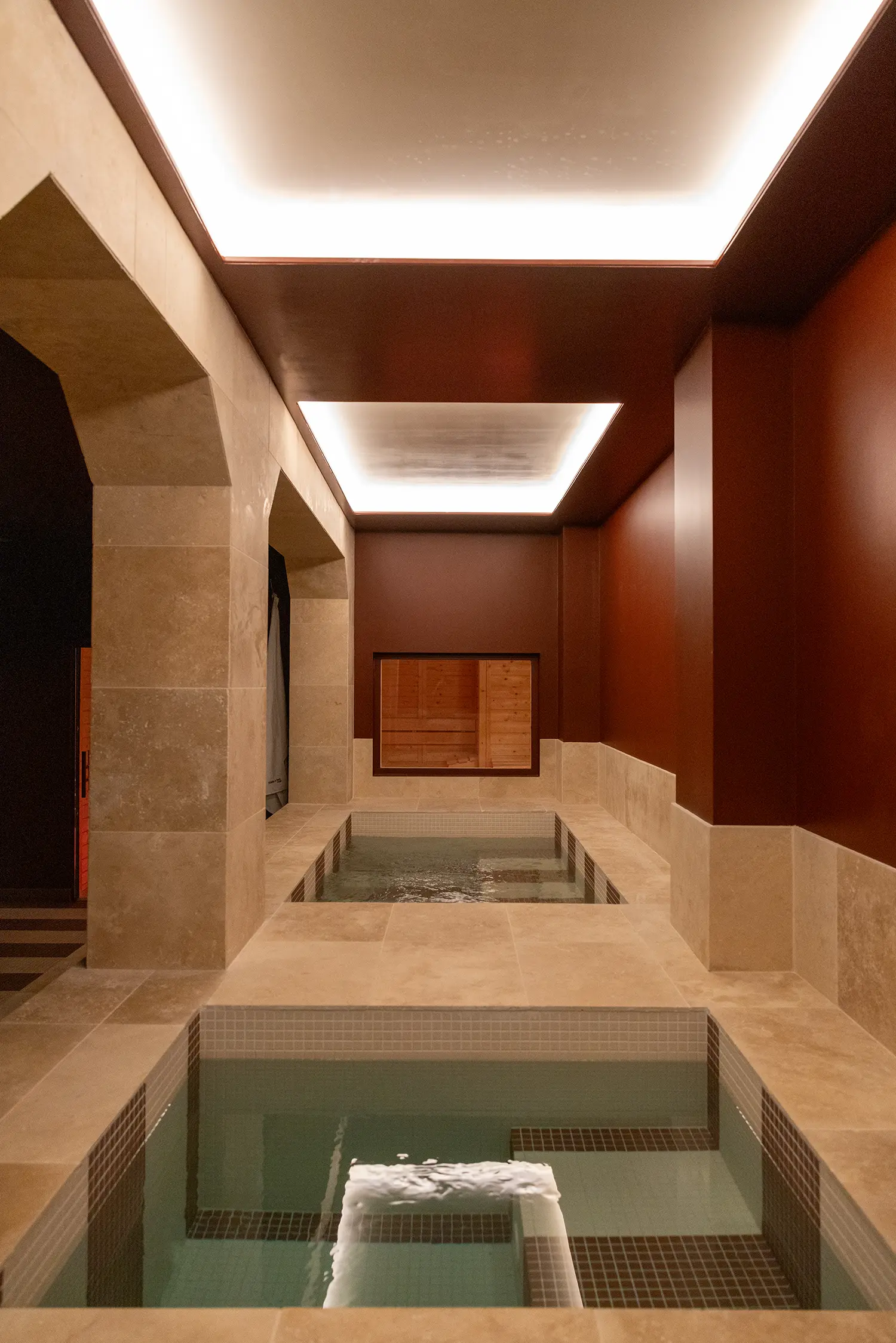 A perspective view of a stone-clad wellness area with square thermal baths and recessed ceiling lighting.