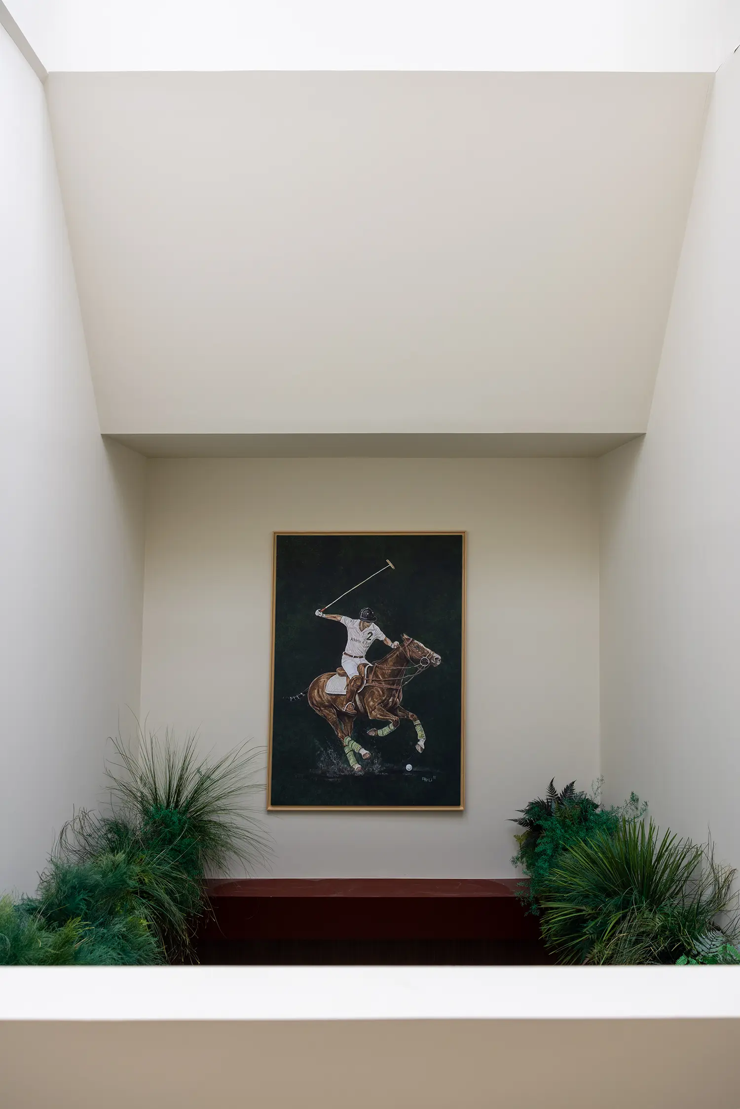 A framed painting of a polo player on a horse centered in a white recessed lightwell surrounded by green plants.