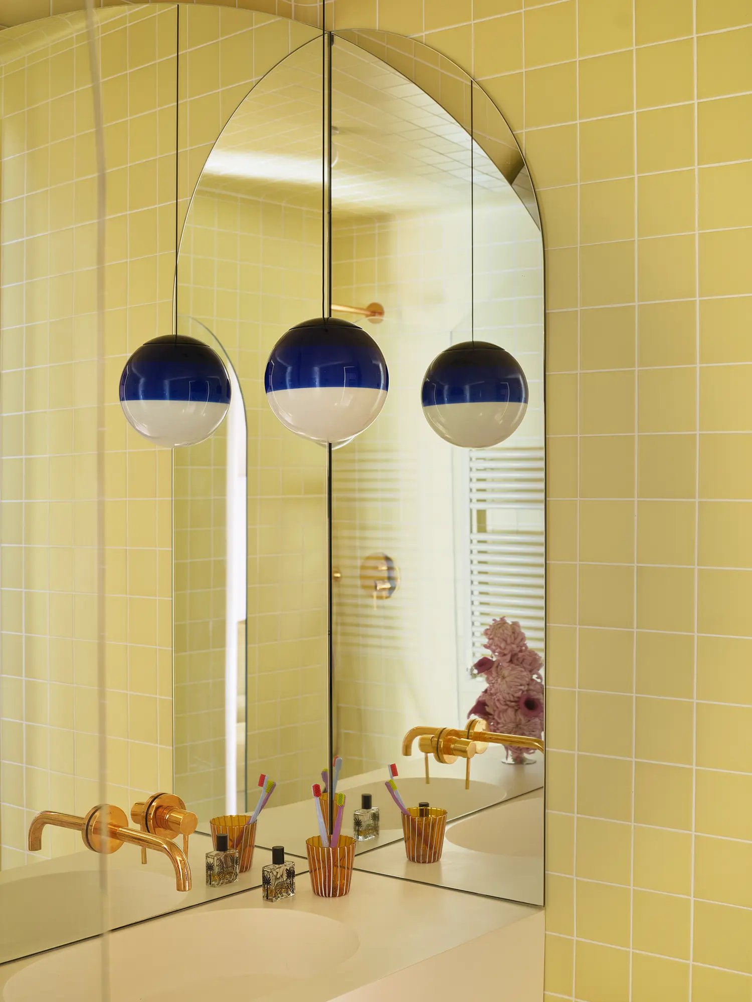 Close-up of a yellow tiled bathroom with an arched mirror, gold faucets, and blue and white spherical pendant lights.