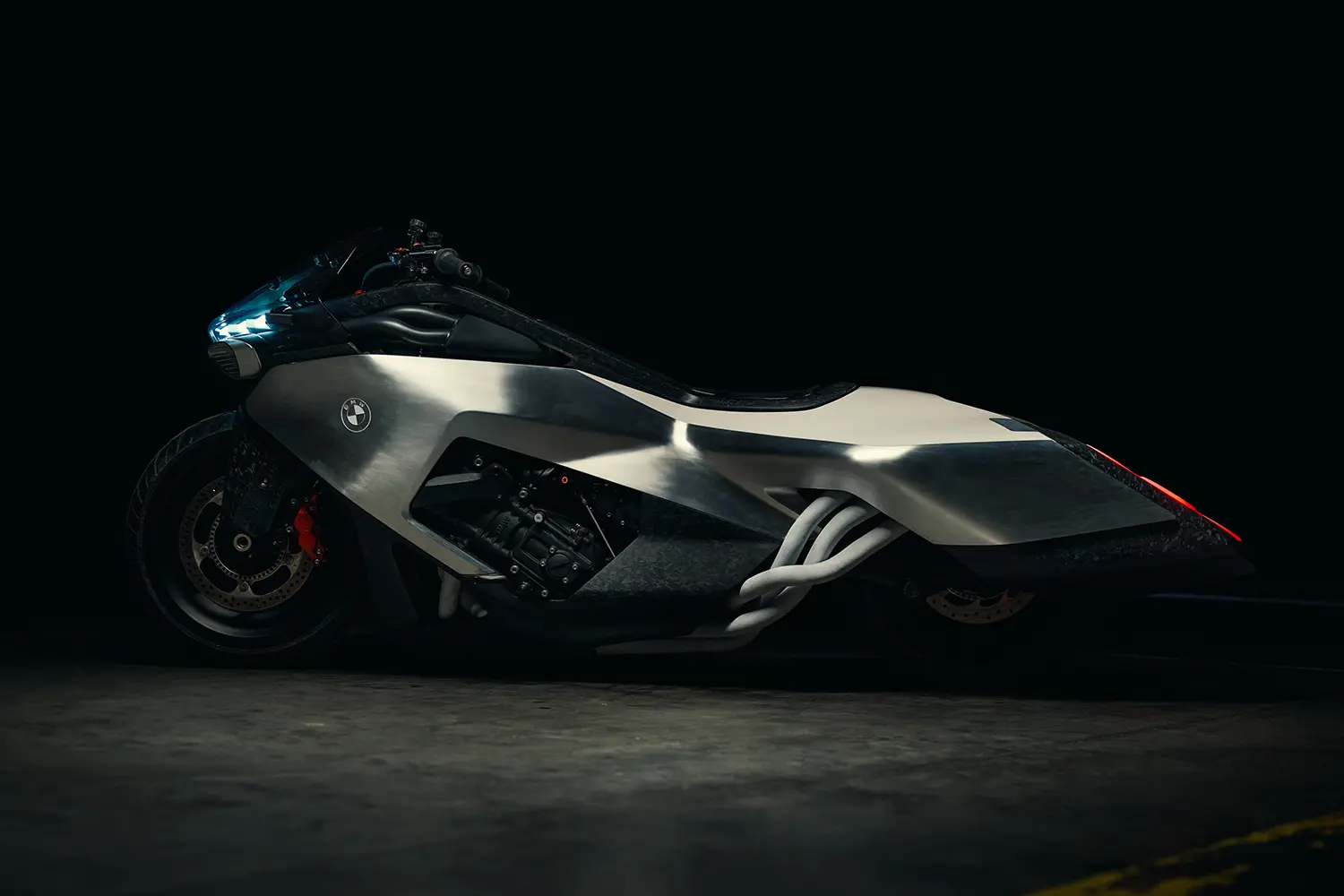 A clean, studio-style side profile of the silver and forged carbon BMW Motorrad Vision K18 against a seamless dark background, with glowing blue headlights and a hint of the tail structure.