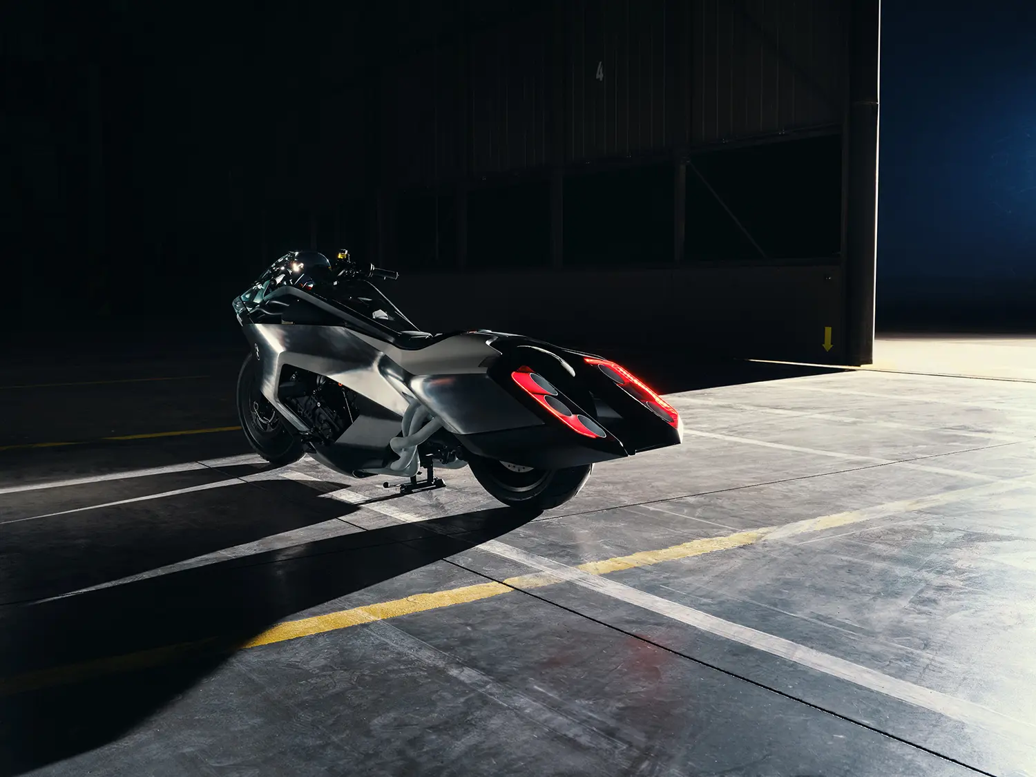 Wide-angle view from a front-quarter perspective, looking across a dark industrial floor toward the illuminated BMW Motorrad Vision K18 concept, highlighting the massive hangar doors in the background.