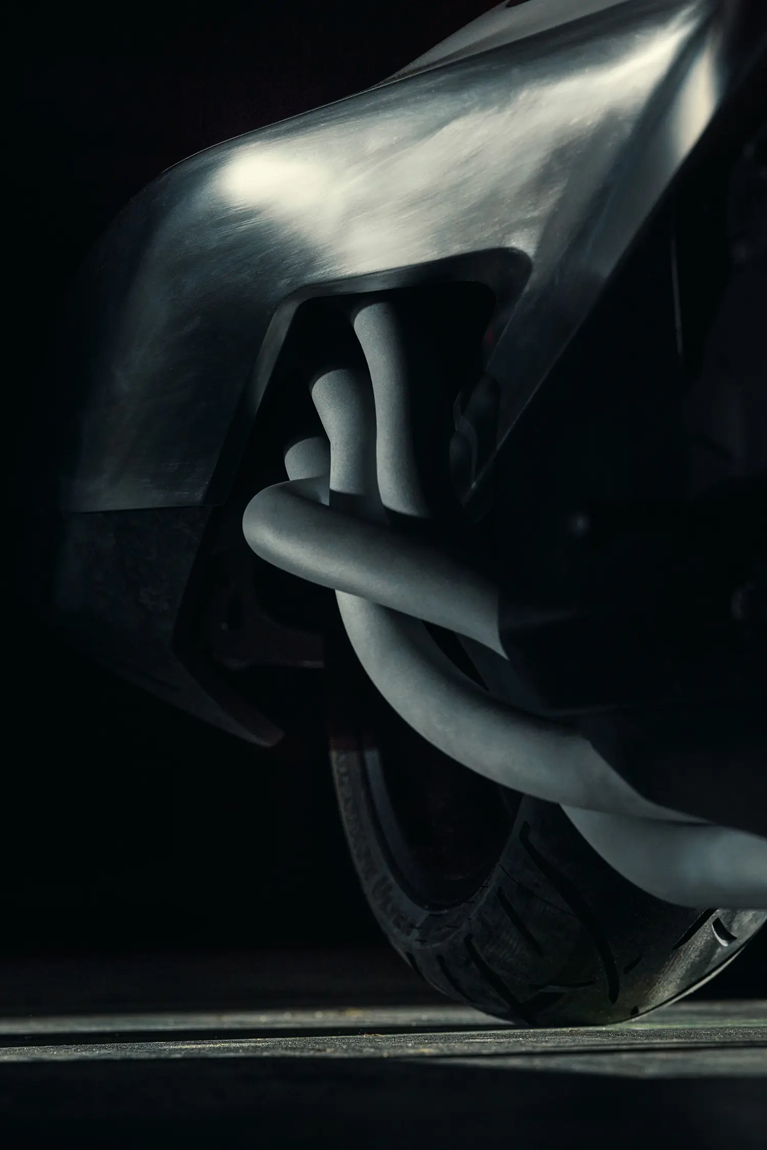 Extreme close-up shot focusing on the complex, intertwined white exhaust headers of the Vision K18 as they exit the engine casting, with a textured tire in the background.