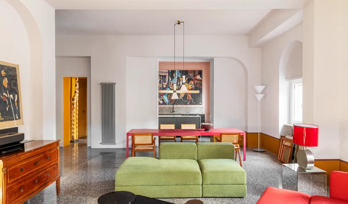 Casa Alkymia by Punto Zero, living and dining area, large-format terrazzo floor, red dining table, green modular sofa, arched counter-wall opening onto pink kitchen, Rome Monteverde