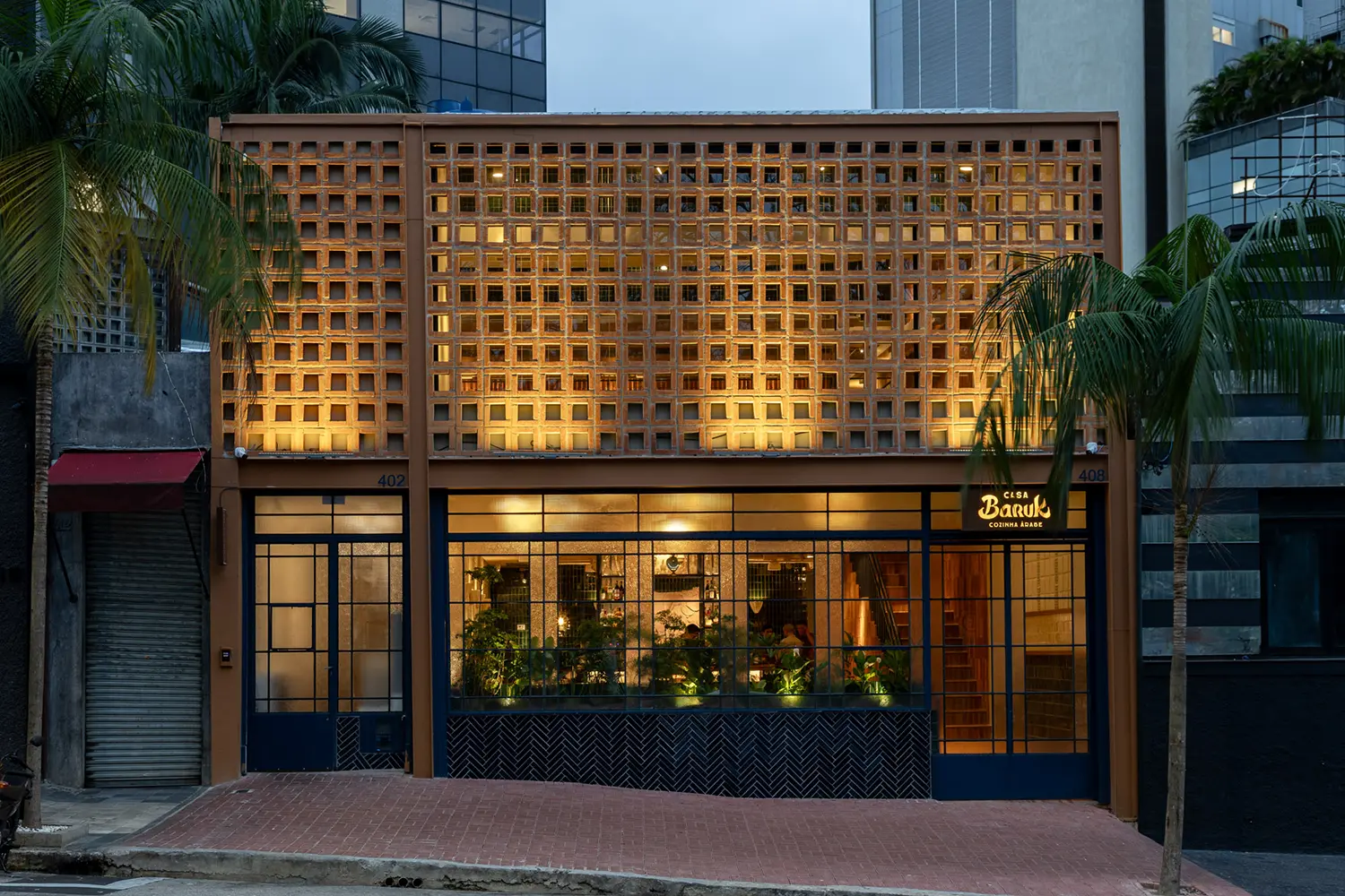 Casa Baruk Restaurant by Memola Estúdio, exterior facade at night, terracotta-painted ceramic Mashrabiya screen, blue iron window casings, illuminated interior visible through glazing, Higienópolis São Paulo