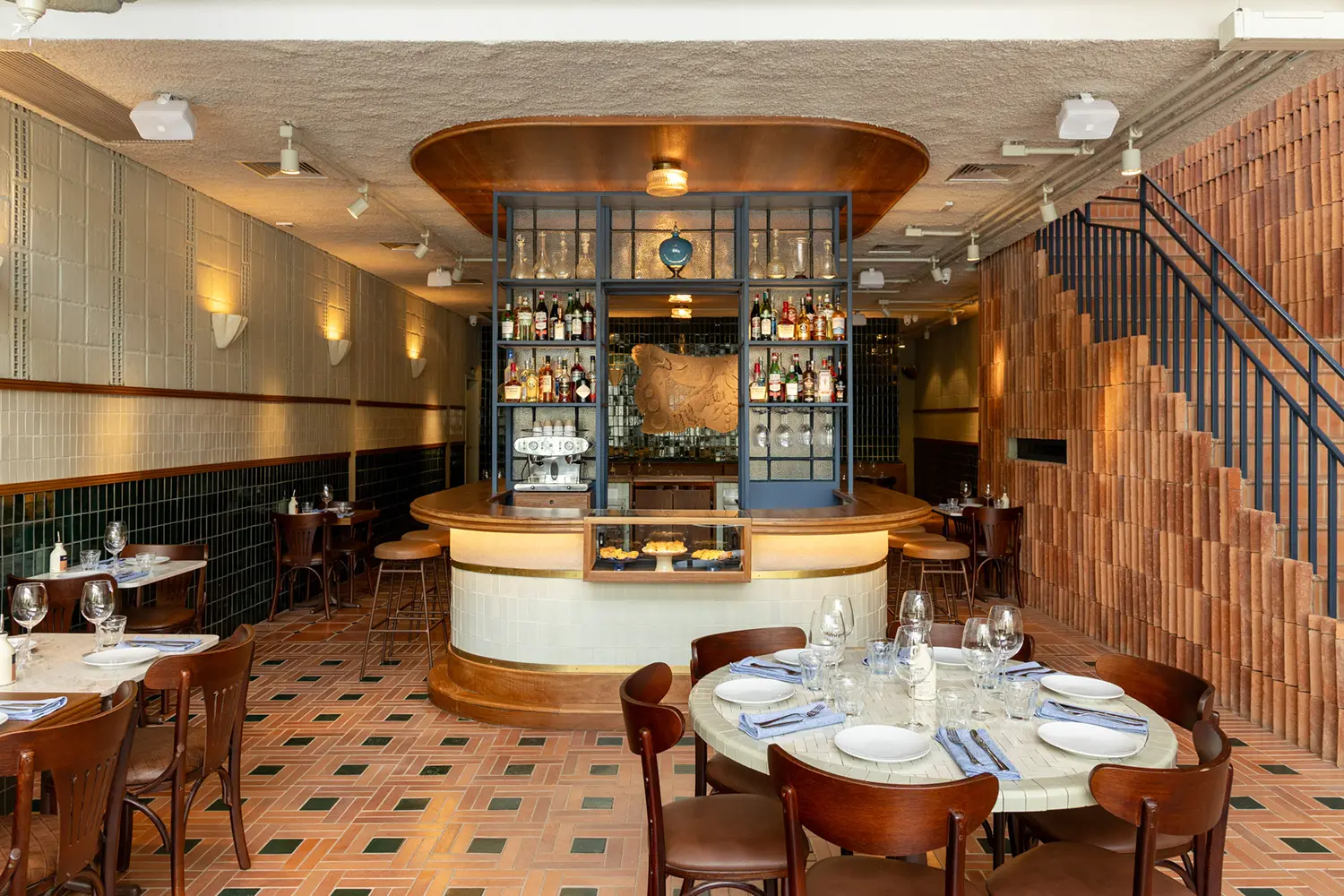Casa Baruk Restaurant by Memola Estúdio, main hall with central bar, rounded ceramic-clad bar base with brass trim, blue iron display cabinet, terracotta and green tile floor, exposed staircase wall, São Paulo