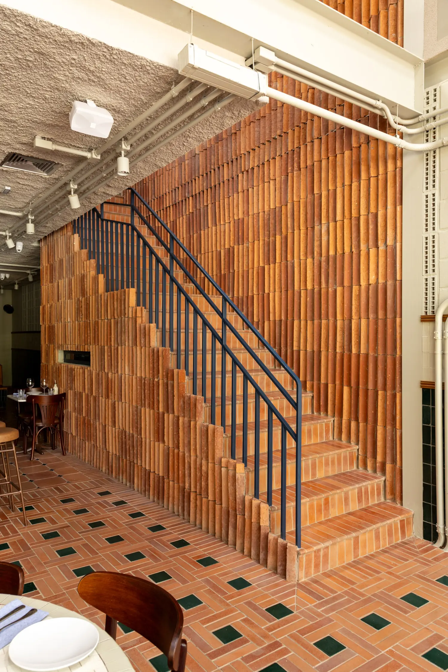 Casa Baruk Restaurant by Memola Estúdio, mezzanine staircase, terracotta ceramic tile wall in vertical stacking pattern, blue iron railing, exposed piping and track lighting, São Paulo