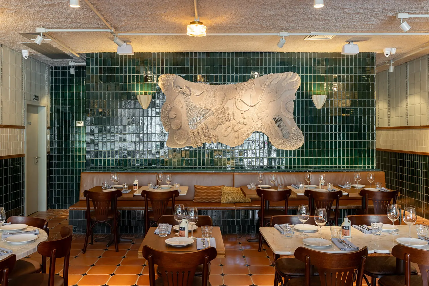 Casa Baruk Restaurant by Memola Estúdio, rear dining area, full-height green ceramic tile back wall, large sculptural white relief artwork, caramel leather banquette, terracotta floor, São Paulo