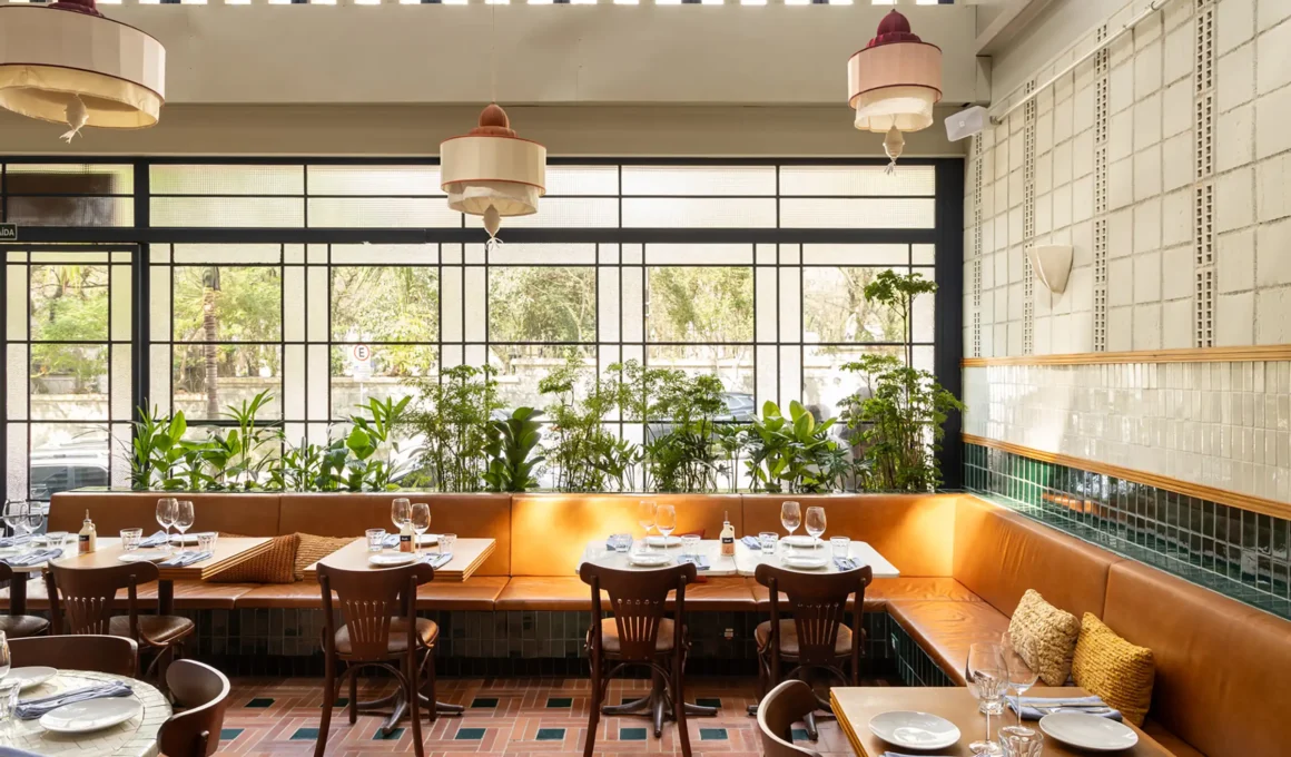 Casa Baruk Restaurant by Memola Estúdio, front dining zone, caramel leather continuous banquette, terracotta and green ceramic tile floor, blue iron window casings, planter strip, custom fabric pendant lights, Higienópolis São Paulo
