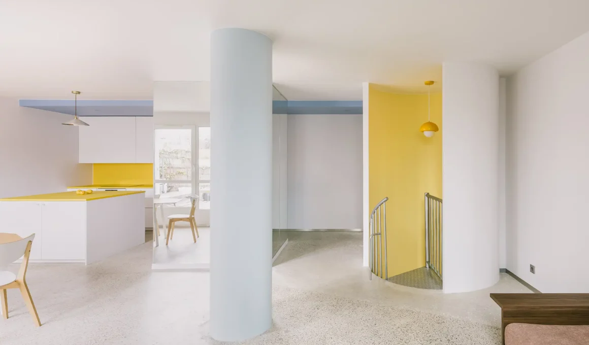 Open-plan living area in a Bagnolet duplex featuring a large light blue pillar, terrazzo flooring, and a yellow staircase niche.