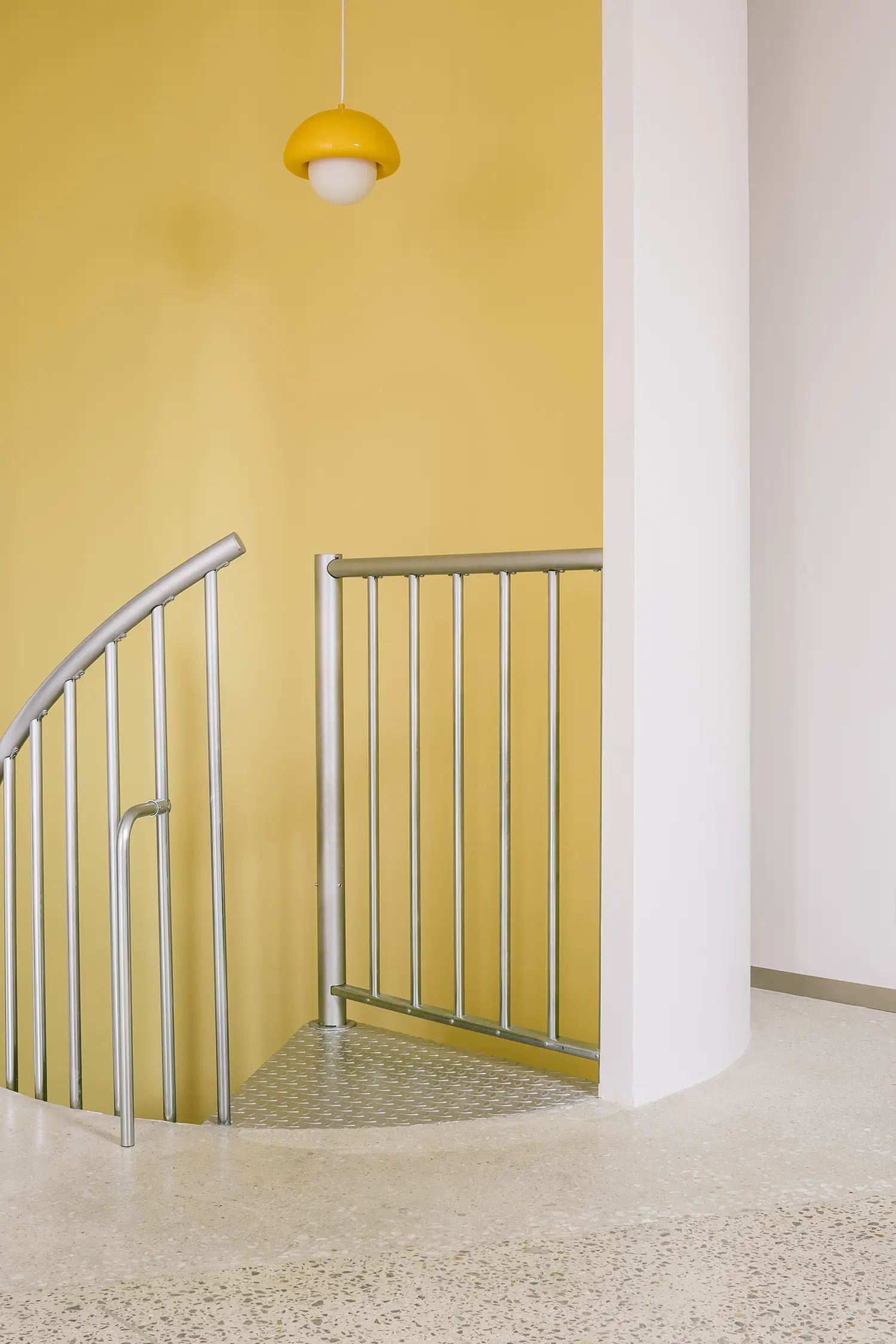 Close-up of a curved yellow staircase wall with a silver metal railing and a matching yellow dome pendant light.