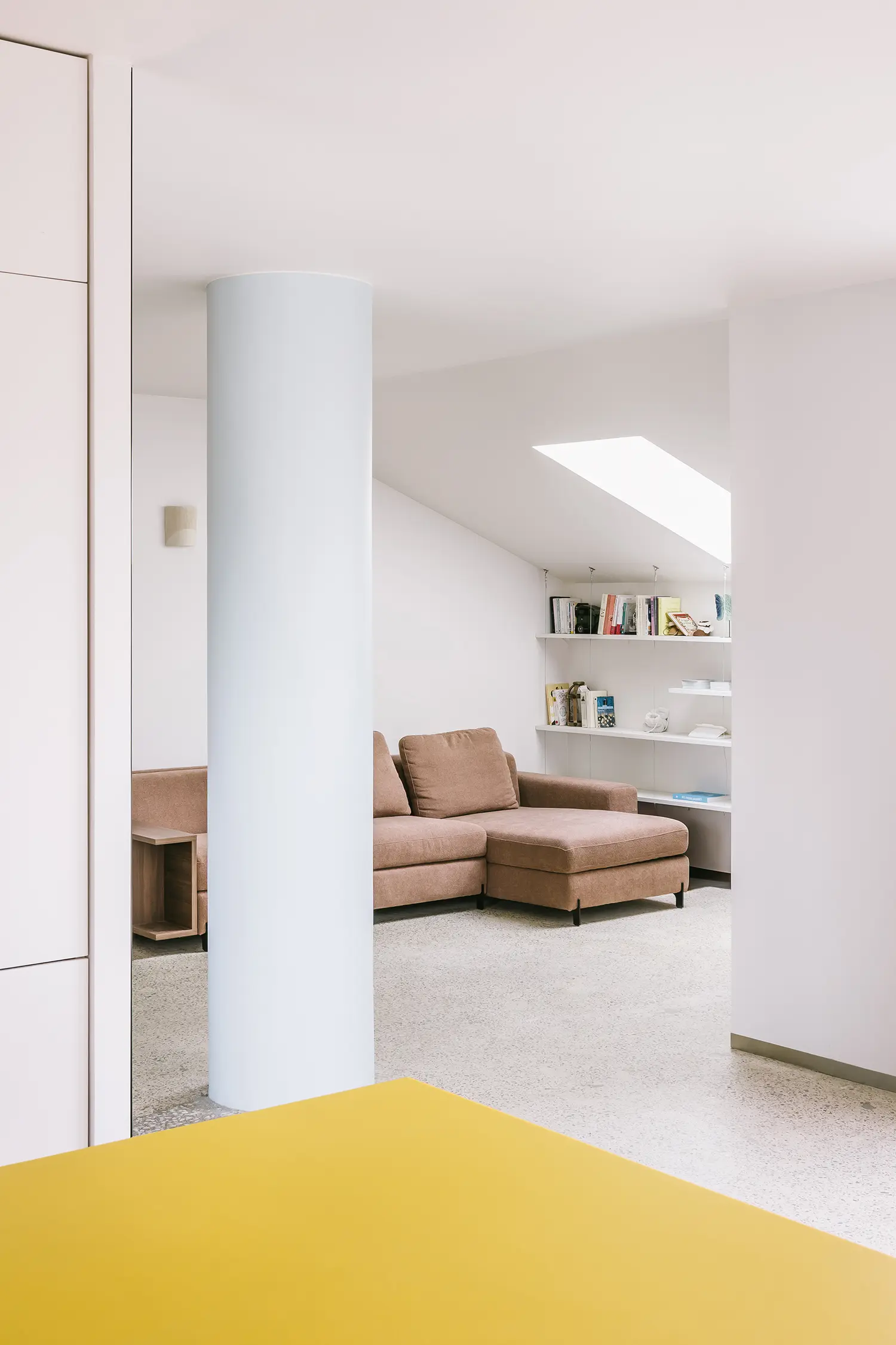 A lounge area with a brown sectional sofa and white built-in shelving, partially obscured by a light blue structural pillar.