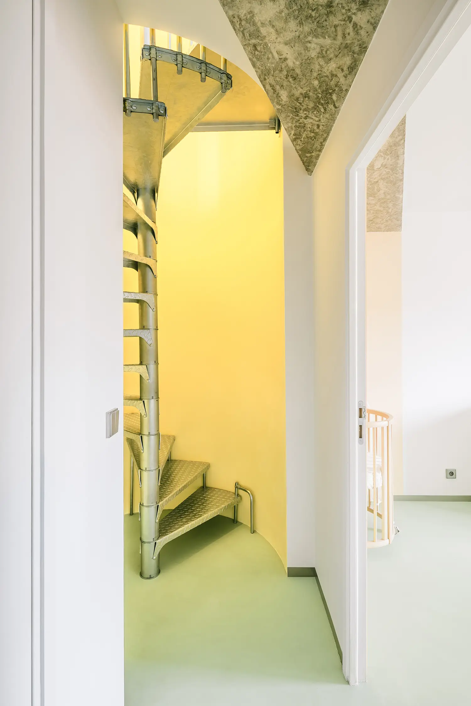 Industrial spiral staircase with silver metal treads set against a bright yellow wall, seen from a doorway.