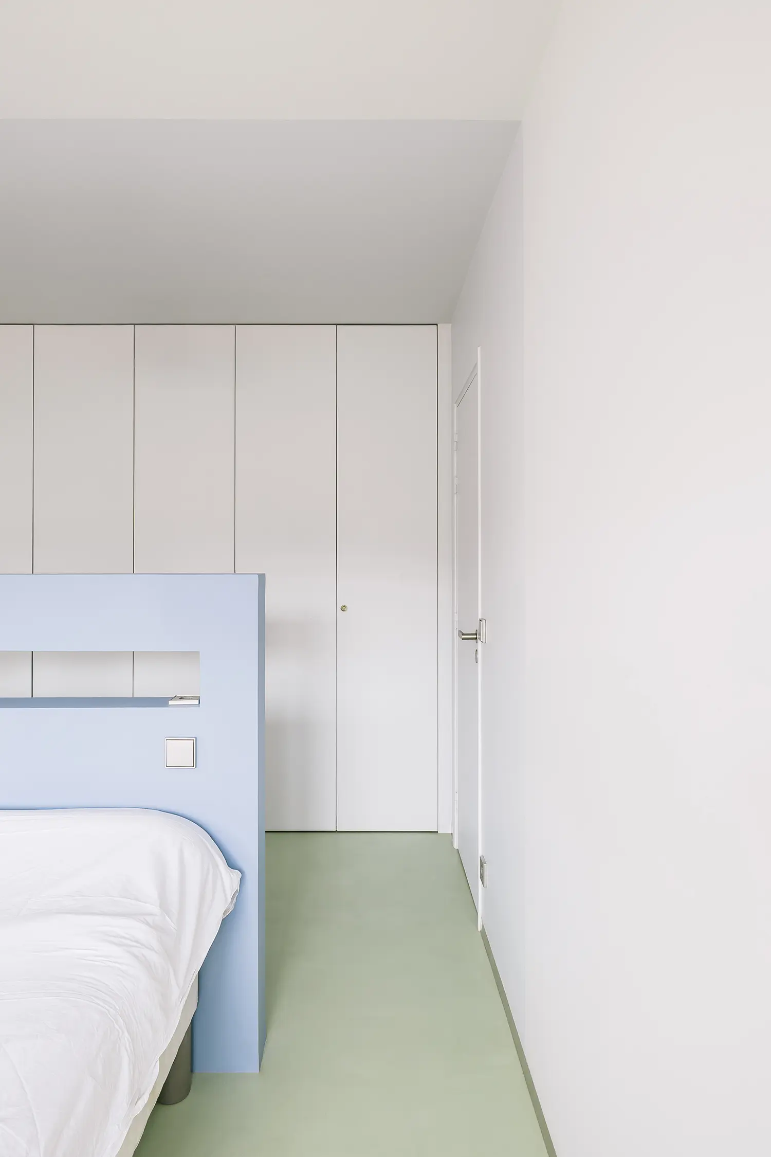 Minimalist bedroom featuring a white bed with a light blue headboard divider and pale green flooring.